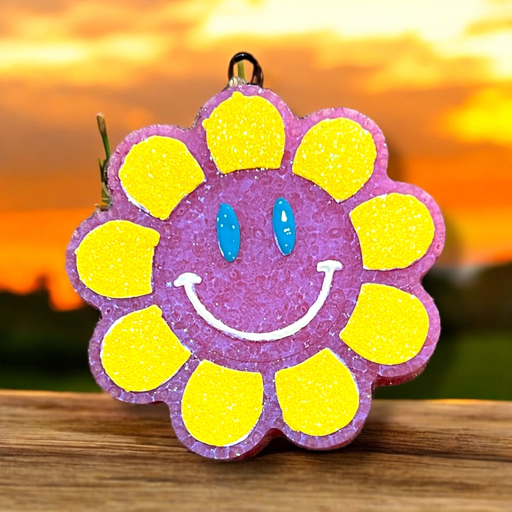 A bright large sunflower-shaped car freshener with glittered petals and a smiling face in the center, displayed against a sunset background.