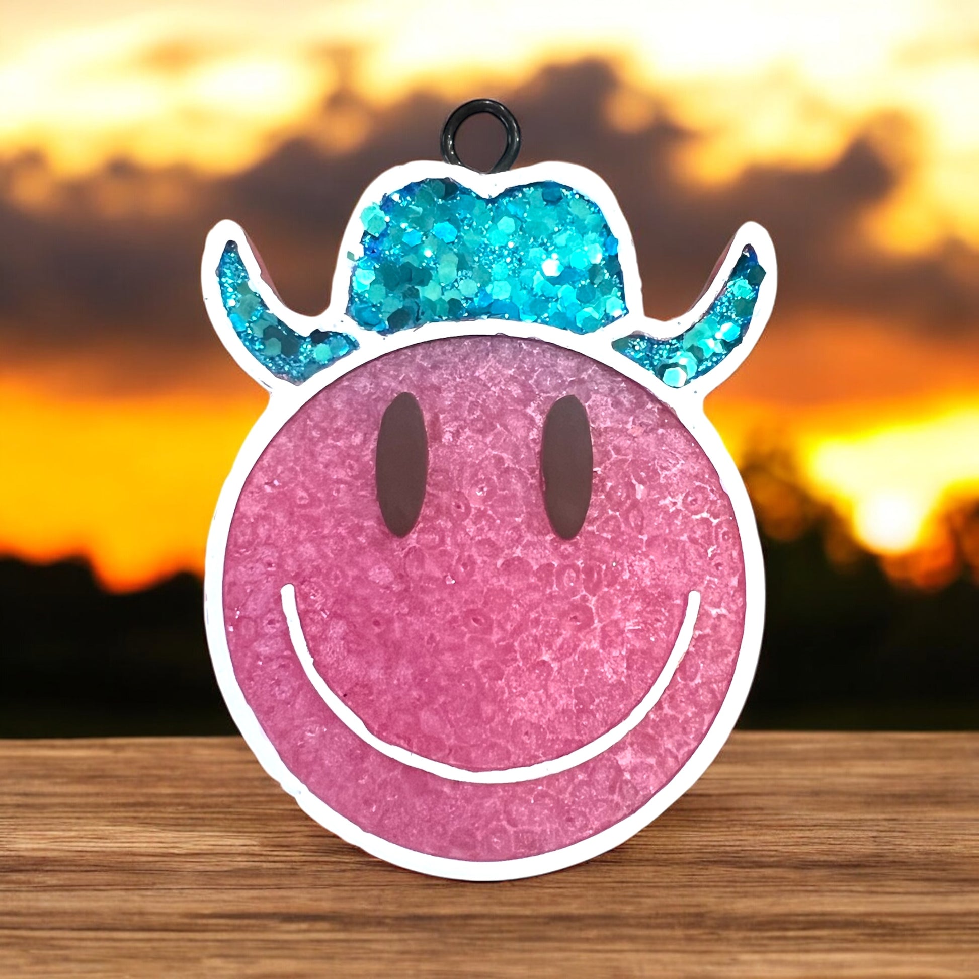 Pink smiley face with blue glittery horns on a wooden surface with a sunset background