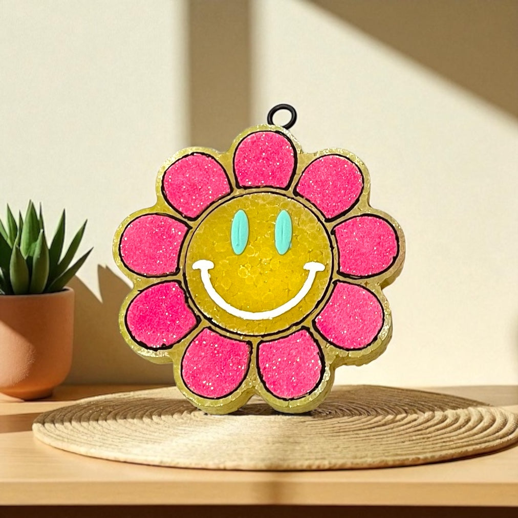 Decorative yellow and pink sunflower car freshie with a smiley face design on a wooden surface with a plant in the background