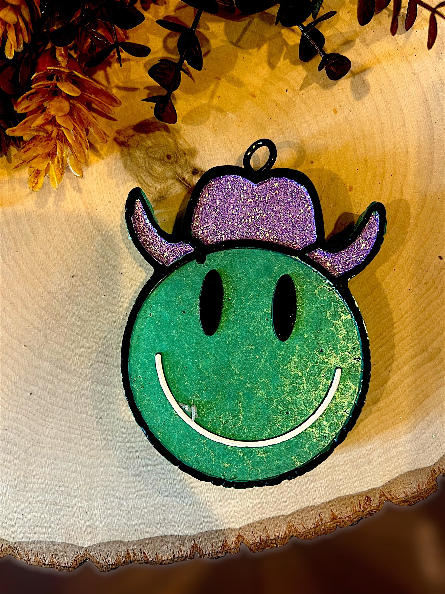 Green smiley face western car freshie with purple hat on a wooden surface with flowers