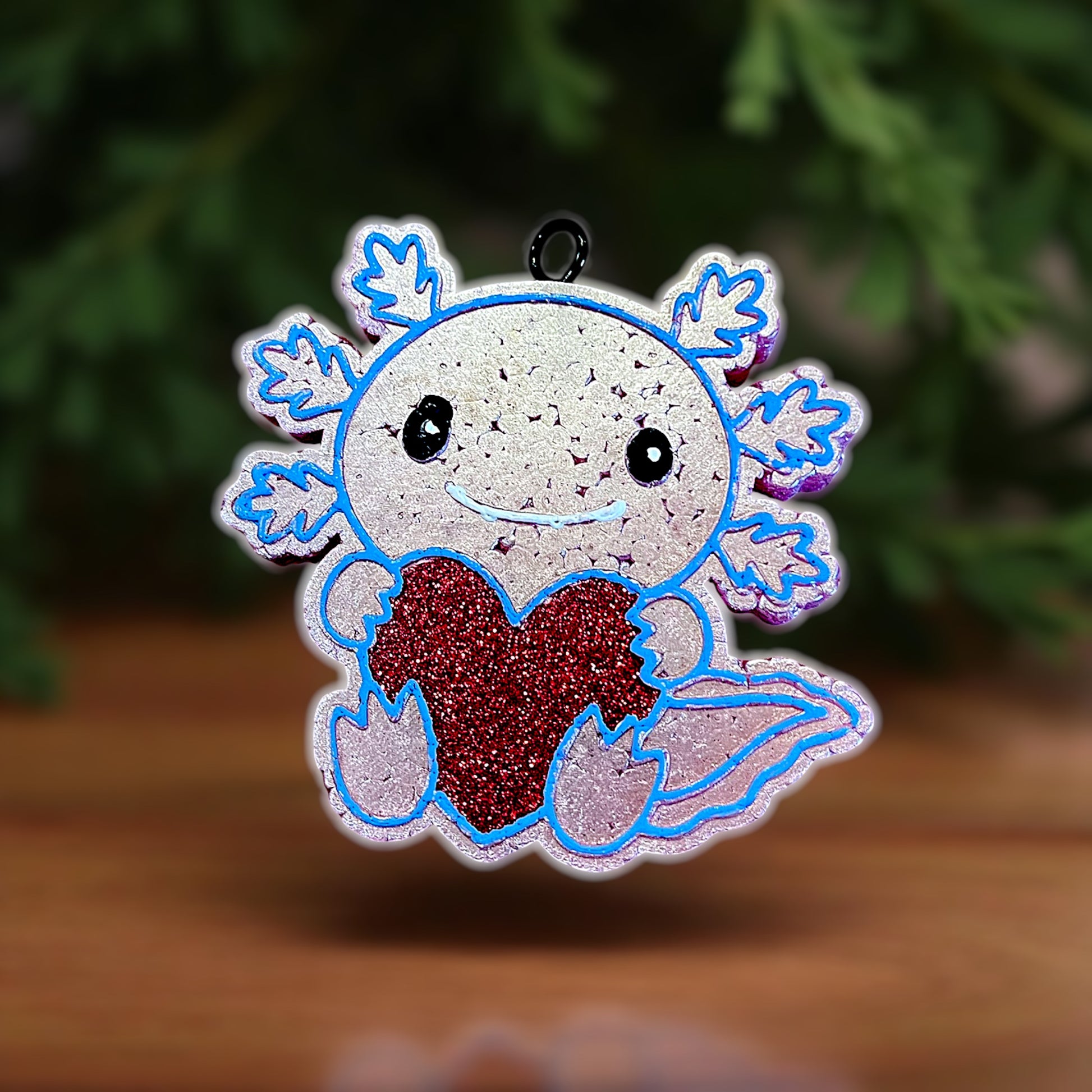 Decorative light pink car freshie with a cartoon axolotl holding a red heart against a blurred green and brown background.