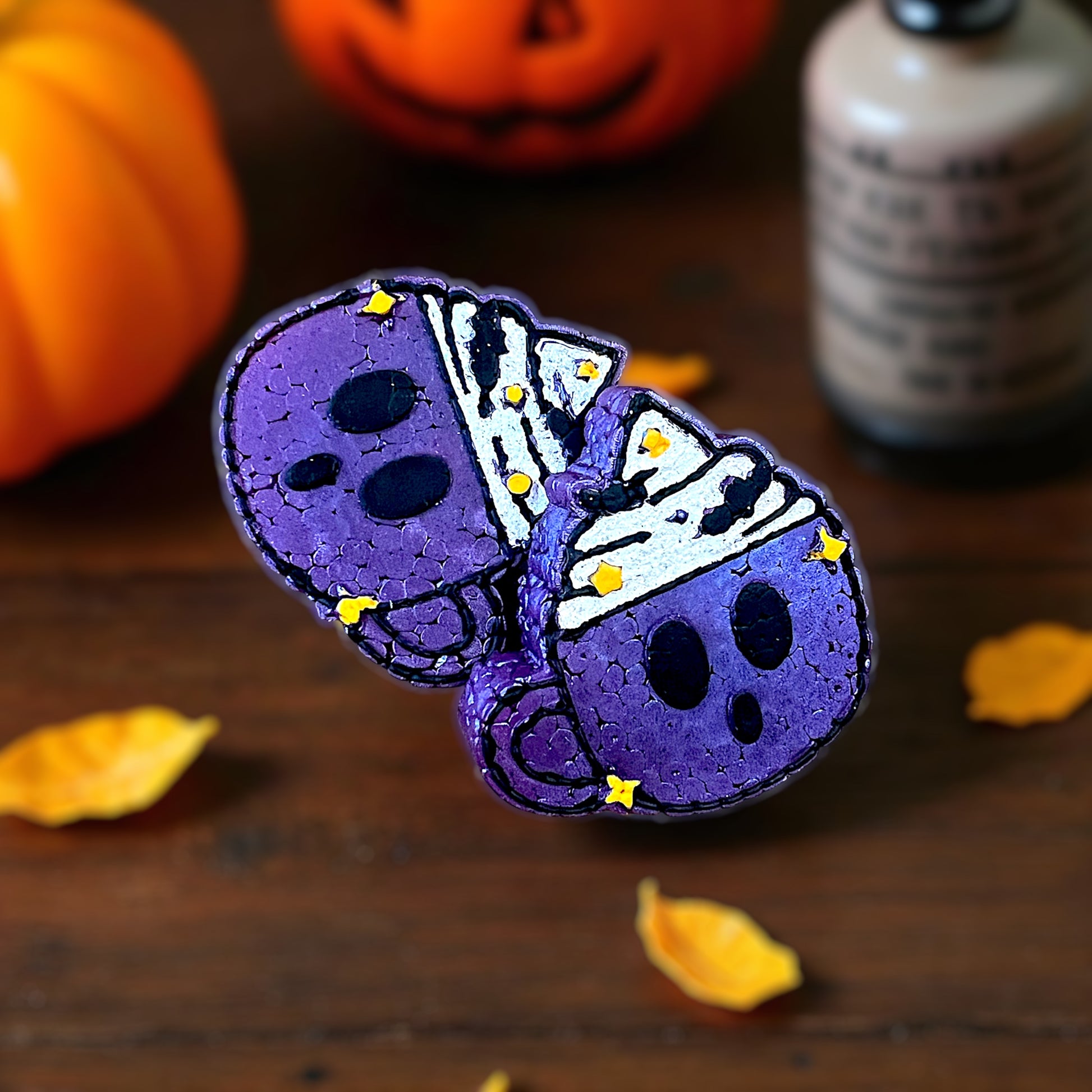 Purple Halloween-themed scented vent clips with black eyes and glittered whipped cream on a wooden surface with pumpkins and leaves.