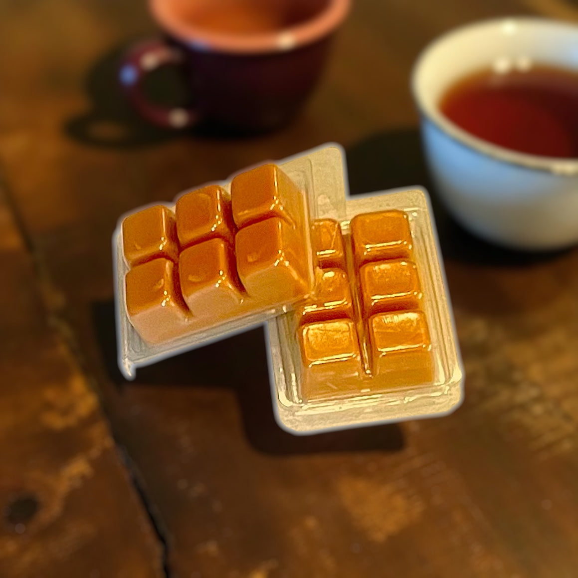 Two containers of orange wax melt clamshells on a wooden surface with two cups in the background.