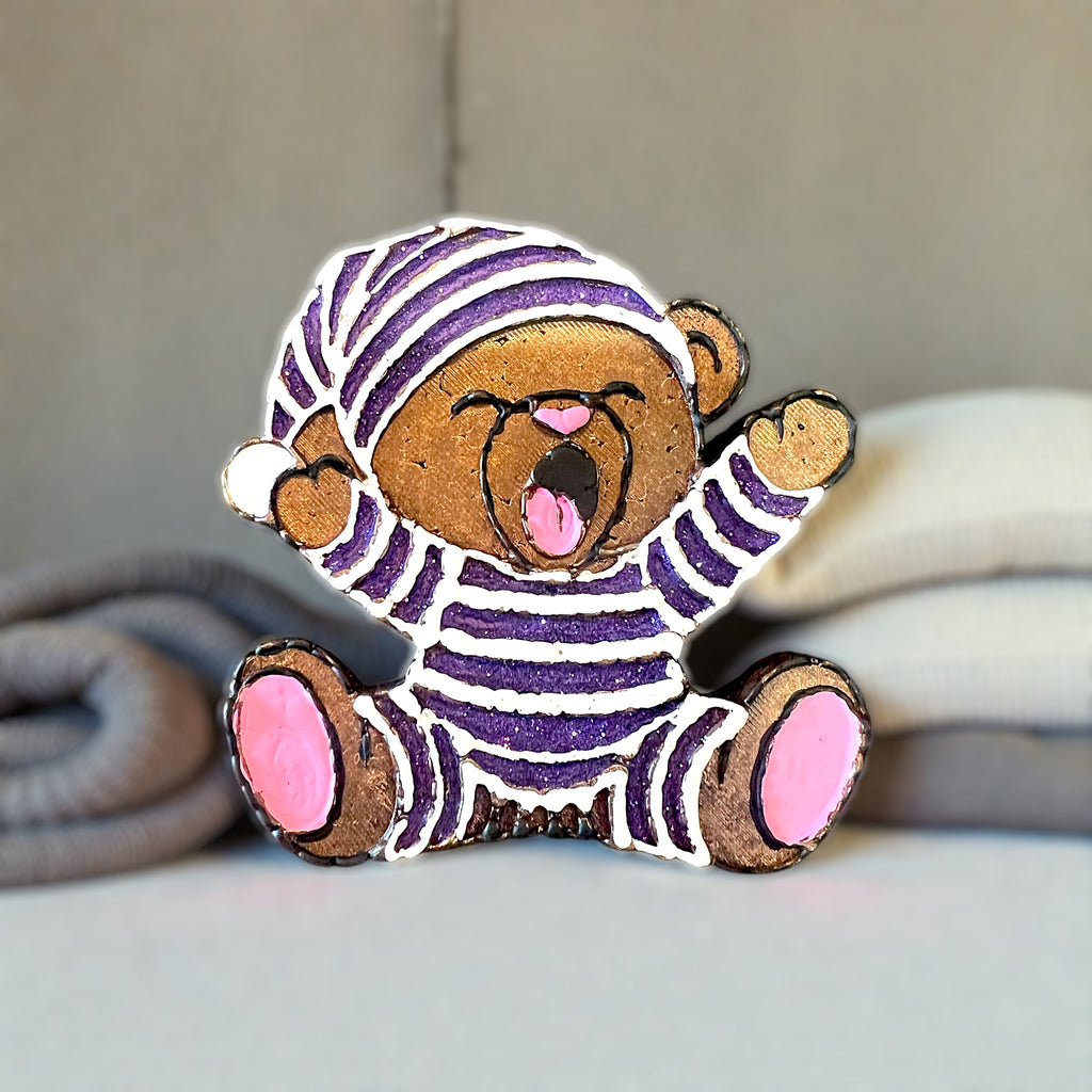 Brown bear car freshie wearing purple glitter pajamas. Yawning and sitting on a bed surrounded by pillows and blankets.