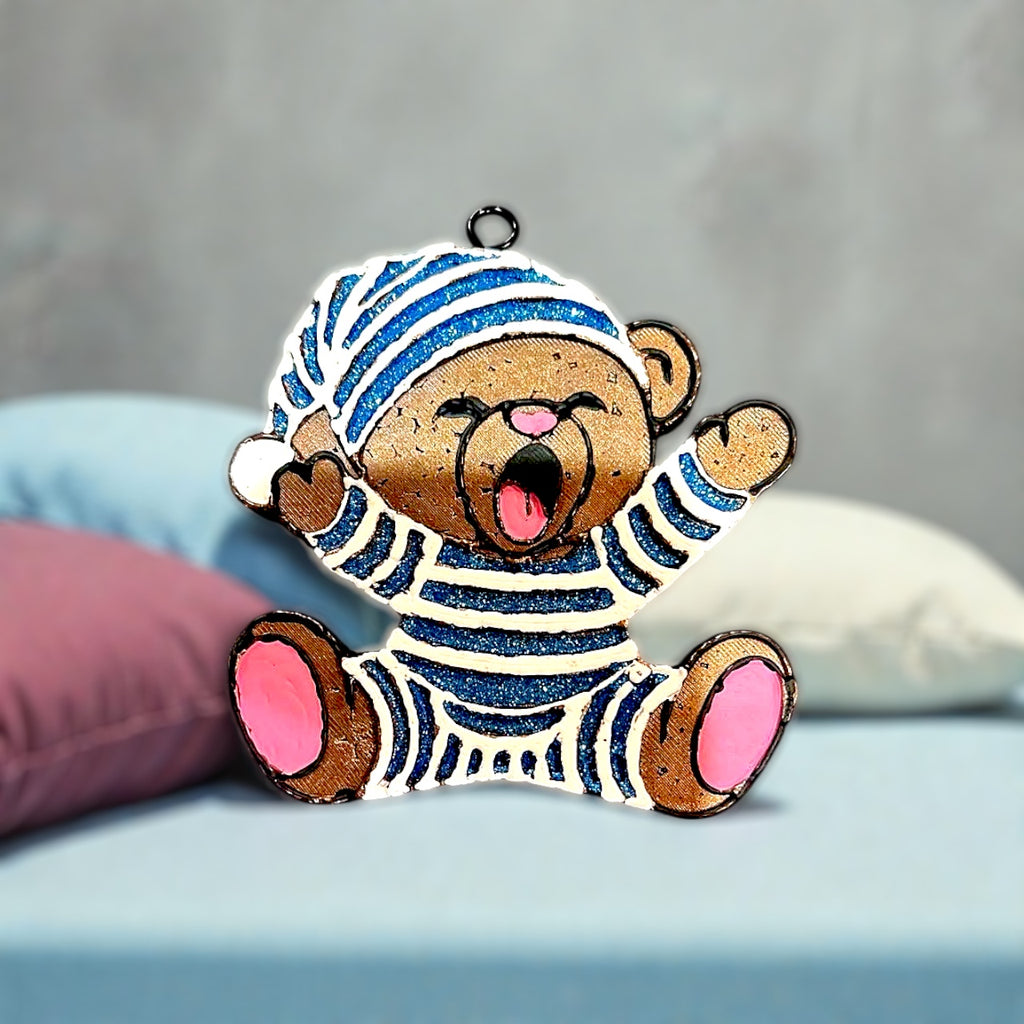 Brown Bear Car Freshie wearing blue glitter pajamas trimmed in white. Yawning and sitting on a bed surrounded by pillows.