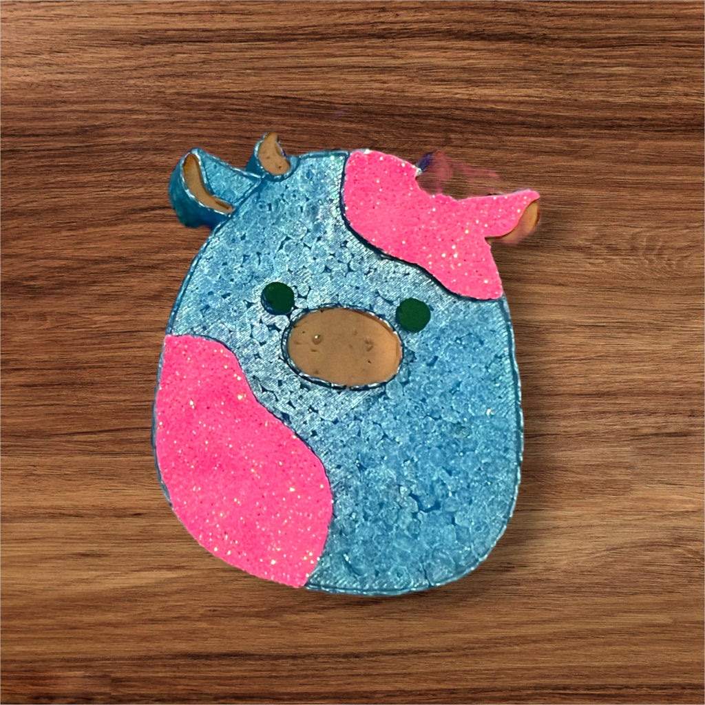 A blue with glittery pink cow-shaped car freshie with a sunset background.