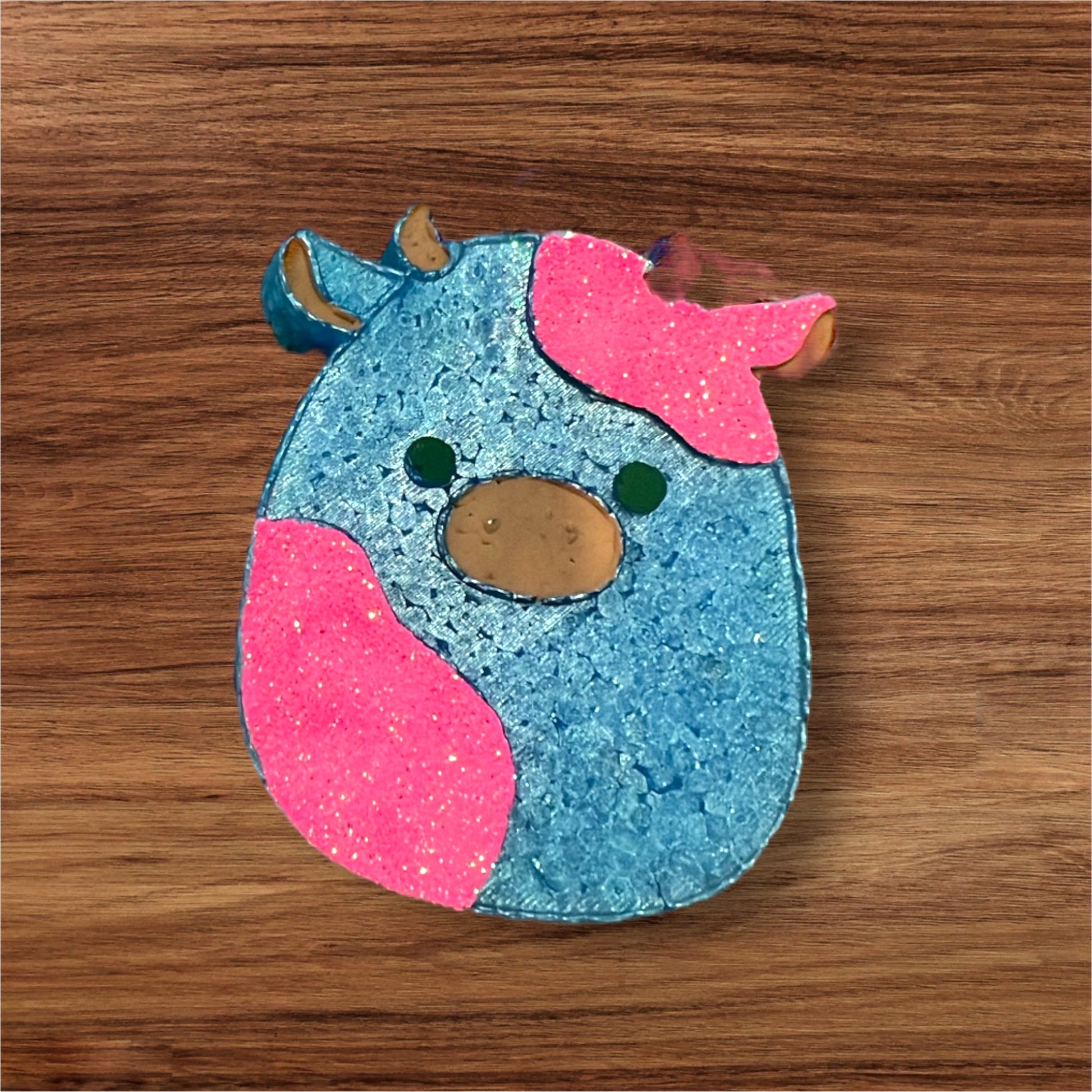 A blue with glittery pink cow-shaped car freshie with a sunset background.