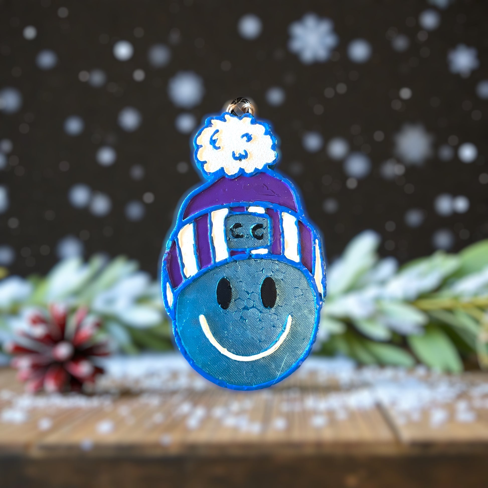 Beanie Waiting for this Season Car Freshie in blue with a purple beanie and a white glitter pom pom on top. Sitting on a wooden table with greenery and snowflakes.