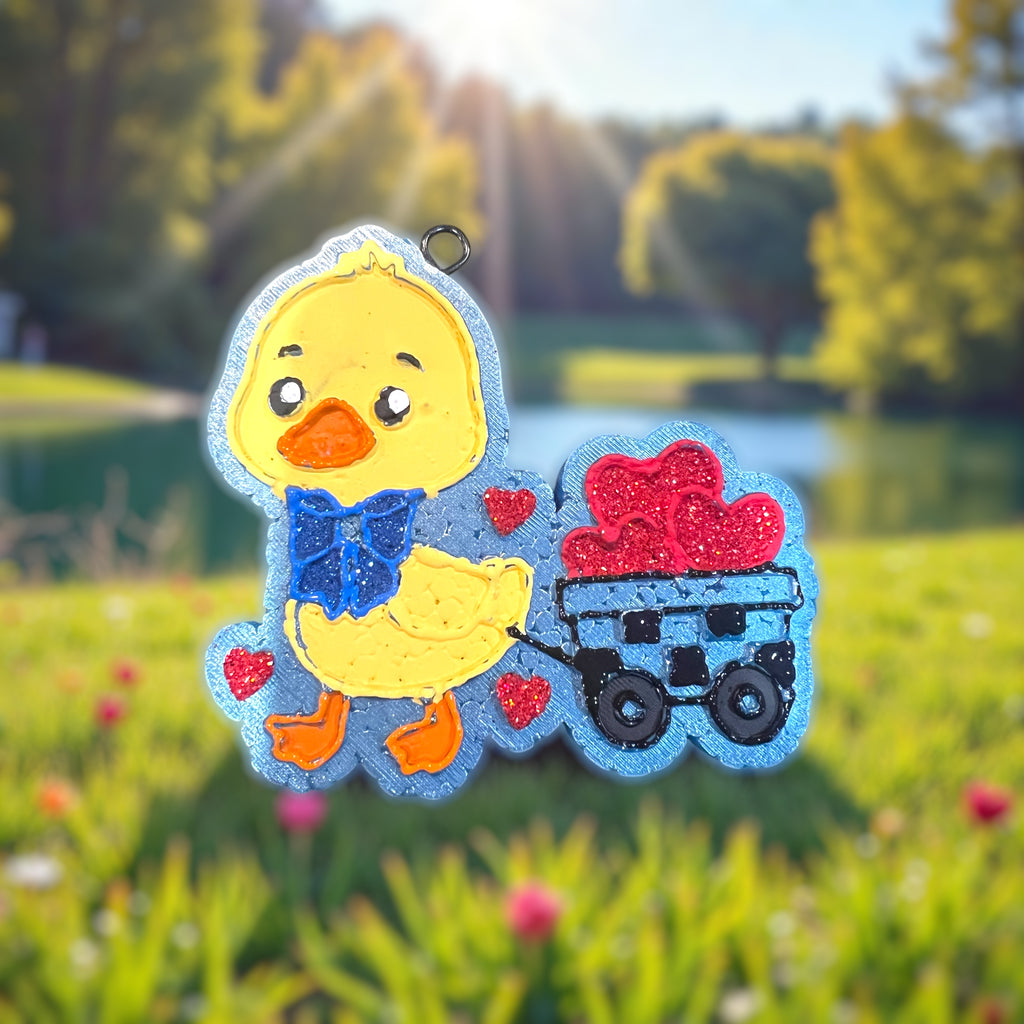 Yellow duck car freshie with a blue glitter bow. Pulling a wagon full of red glitter hearts through the grass. A pond in the background. 