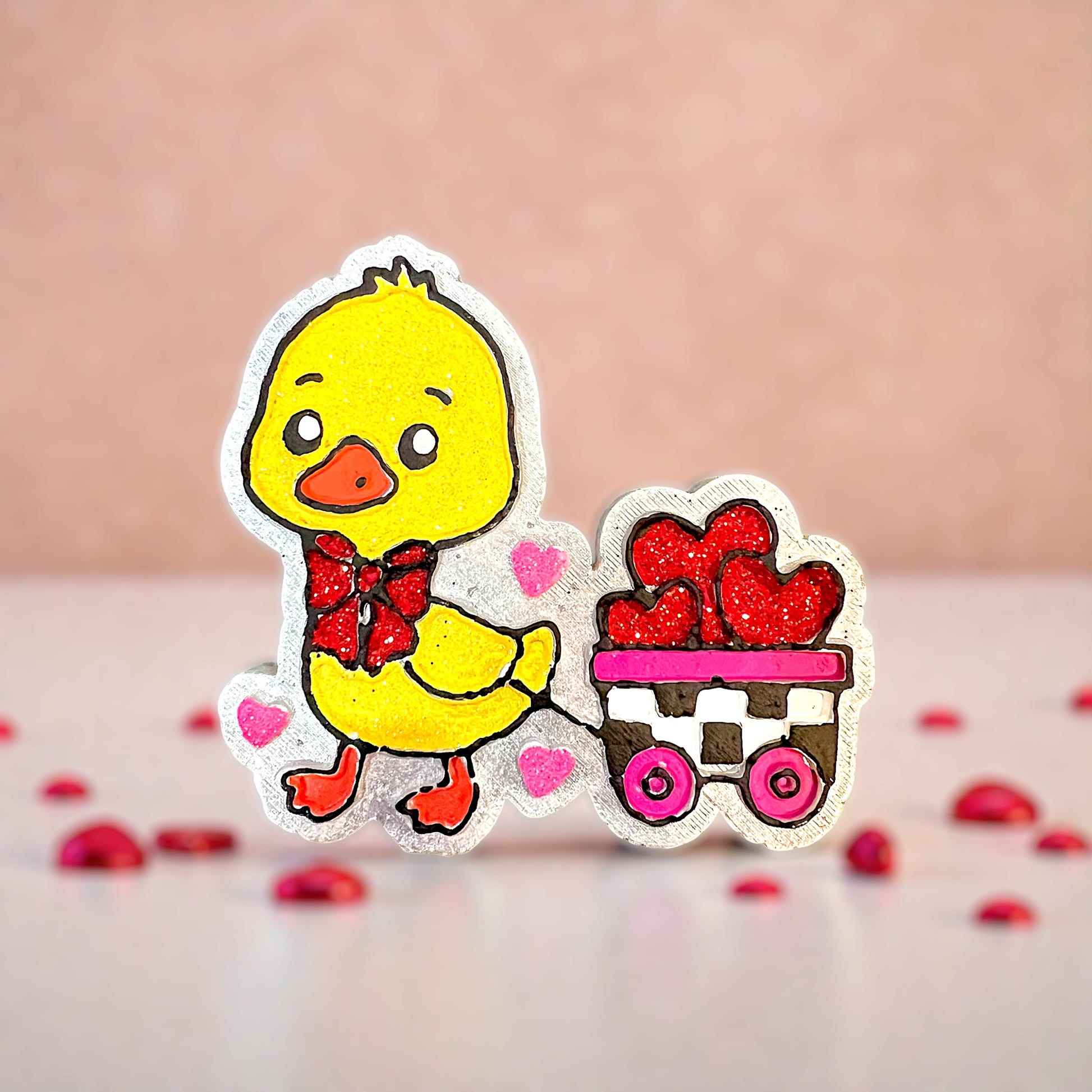 Yellow duck car freshie with a red glittered bow pulling a wagon. Wagon is full of red glittered hearts and trimmed in pink and black. All on a white background.