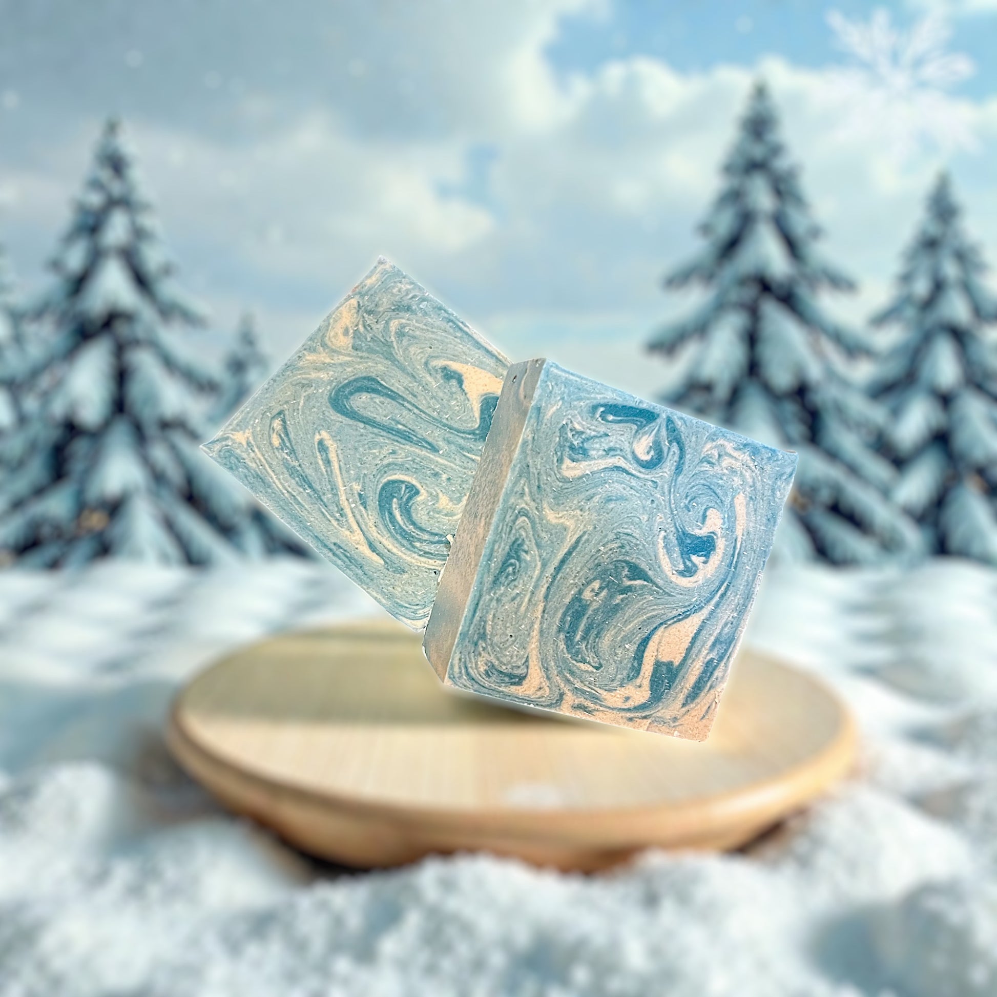 Blue and White swirled goat milk soap sitting on a wooden plate in snow. Background is blurred pine trees covered in snow.