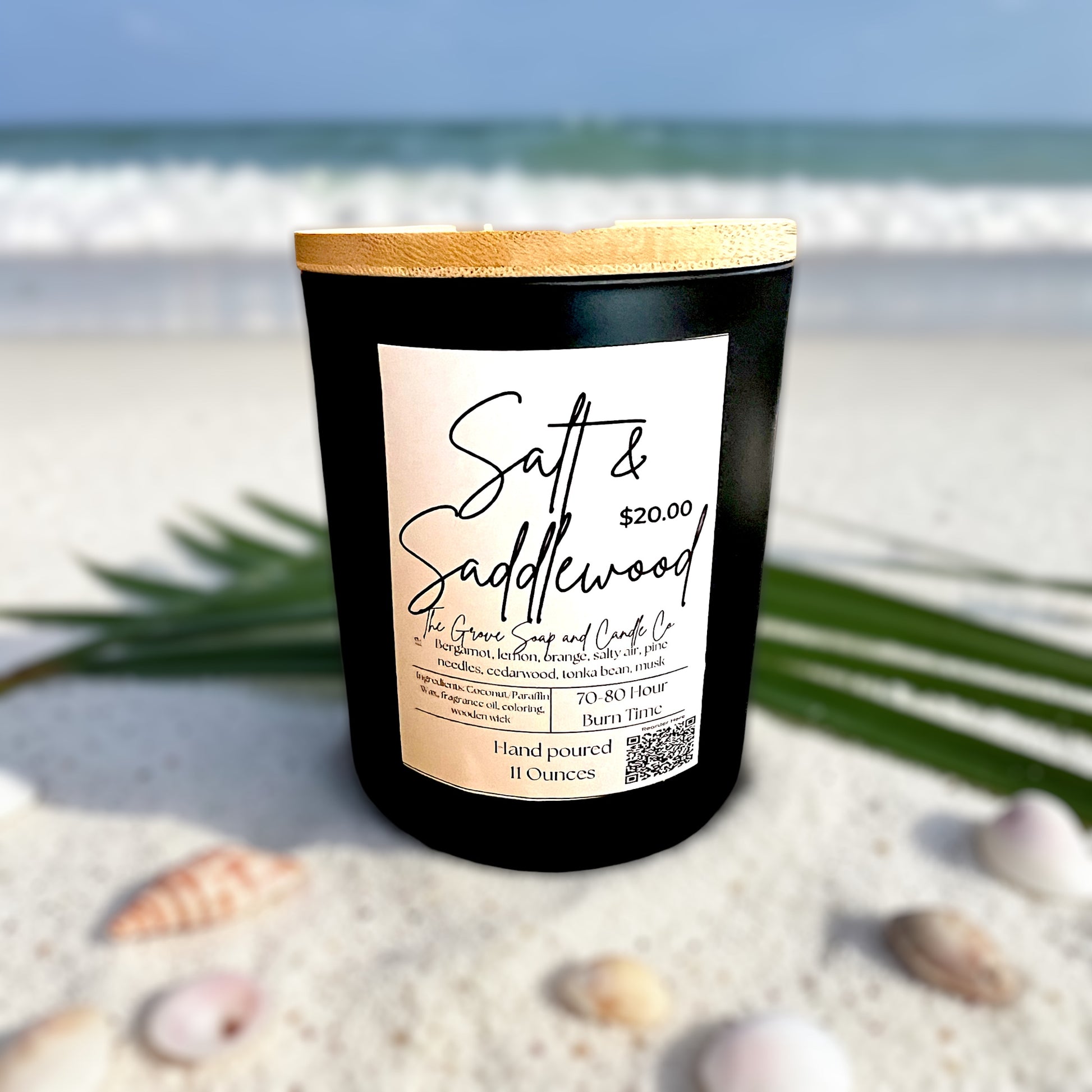 Black Candle jar with a wooden lid. Labeled with the product name Salt & Saddlewood on a sandy beach with ocean in the background