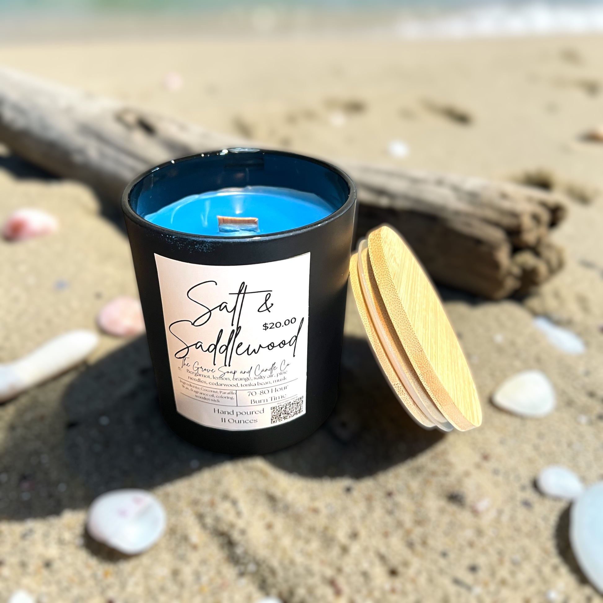 Black candle jar with a wooden lid on a sandy beach. Surrounded by shells and driftwood. Shows the label Salt & Saddlewood and the color blue of the wax. Shows the wooden wick. 