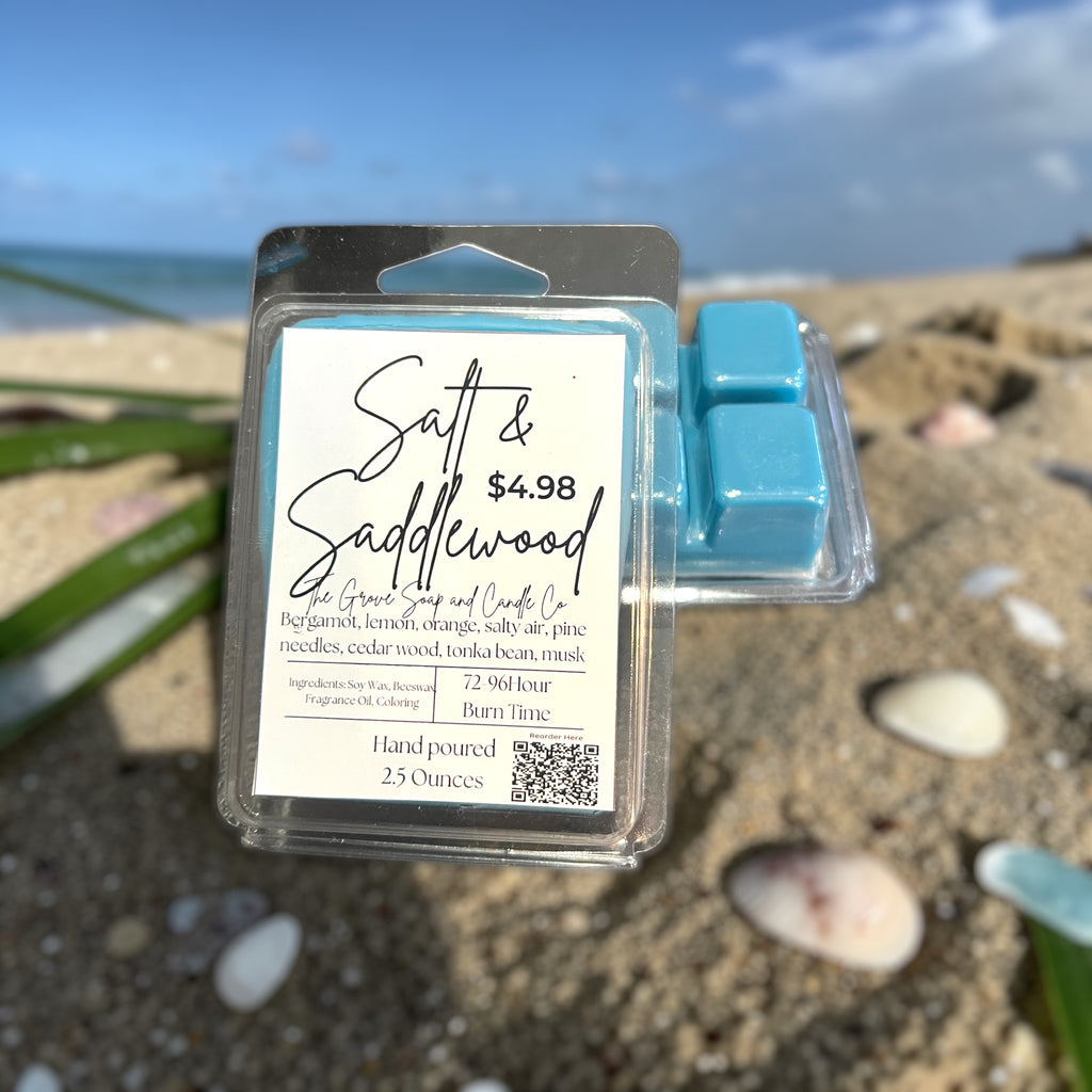 Packaged salt and saddlewood wax melts on a beach with ocean in the background. Shows the label and packaging with ingredients and scent profile. 