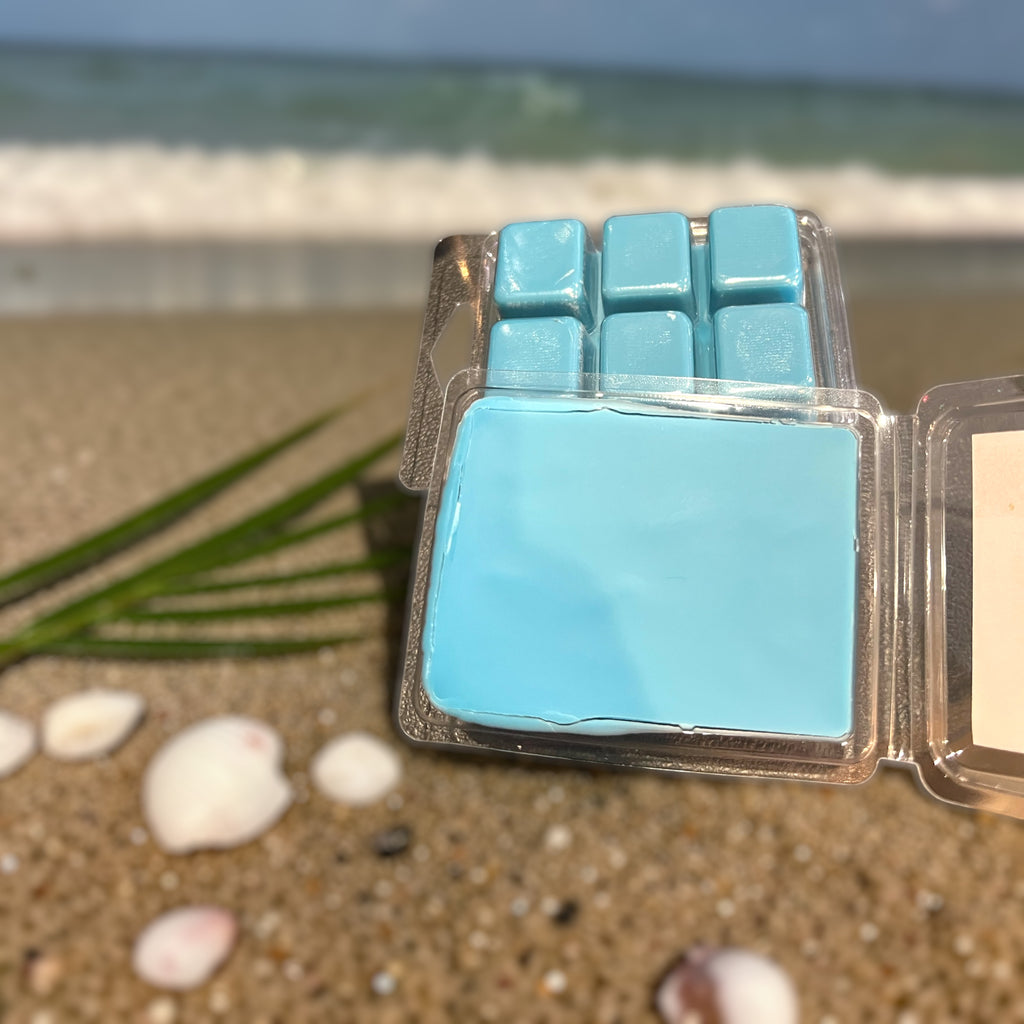 Blue wax melts in a clear container on a sandy beach with ocean in the background