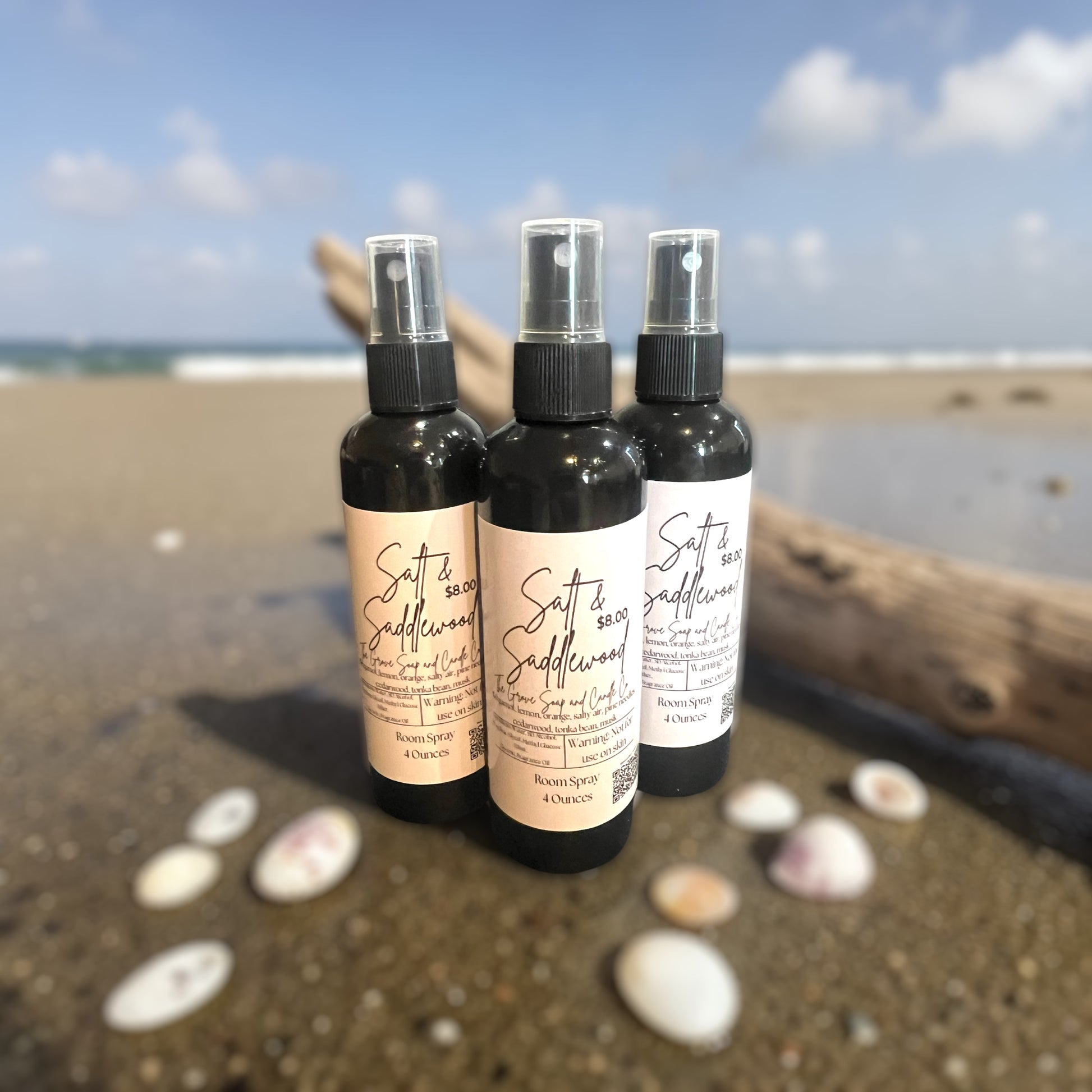 Three black bottles of Salt & Saddlewood Room Spray. Shows the packaging and label with the scent profile. Sitting on the sand at a beach. 