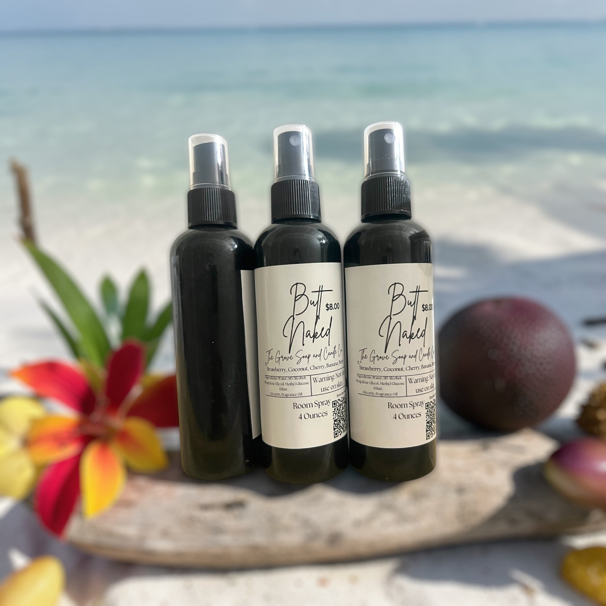 Three bottles of Butt Naked Room Spray sitting on a piece of driftwood on a white sandy beach. Scented in strawberries, cherries, banana, and coconut.