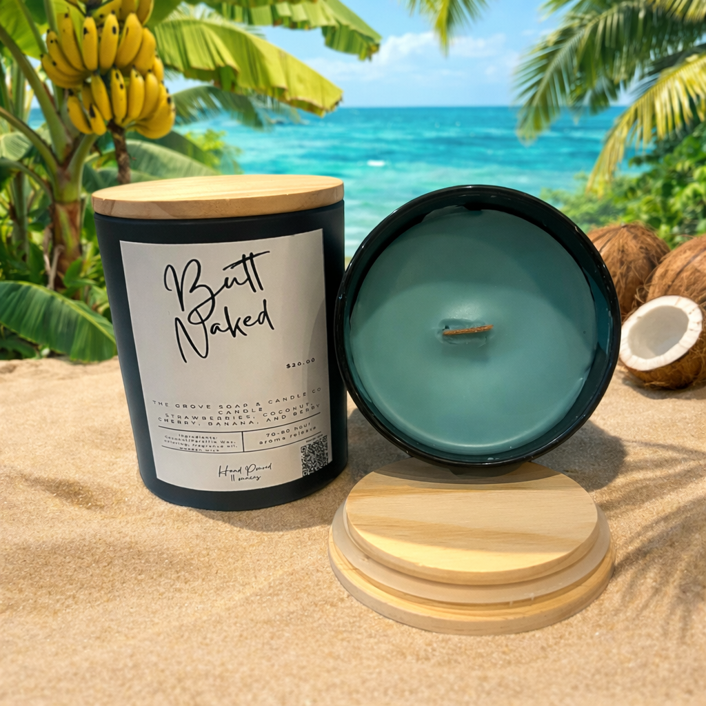 Butt Naked hand poured tropical candle with strawberry, coconut, banana, and cherry fragrance in a wooden wick candle jar by The Grove Soap and Candle Co. sitting on a sandy beach with tropical trees in the background. 