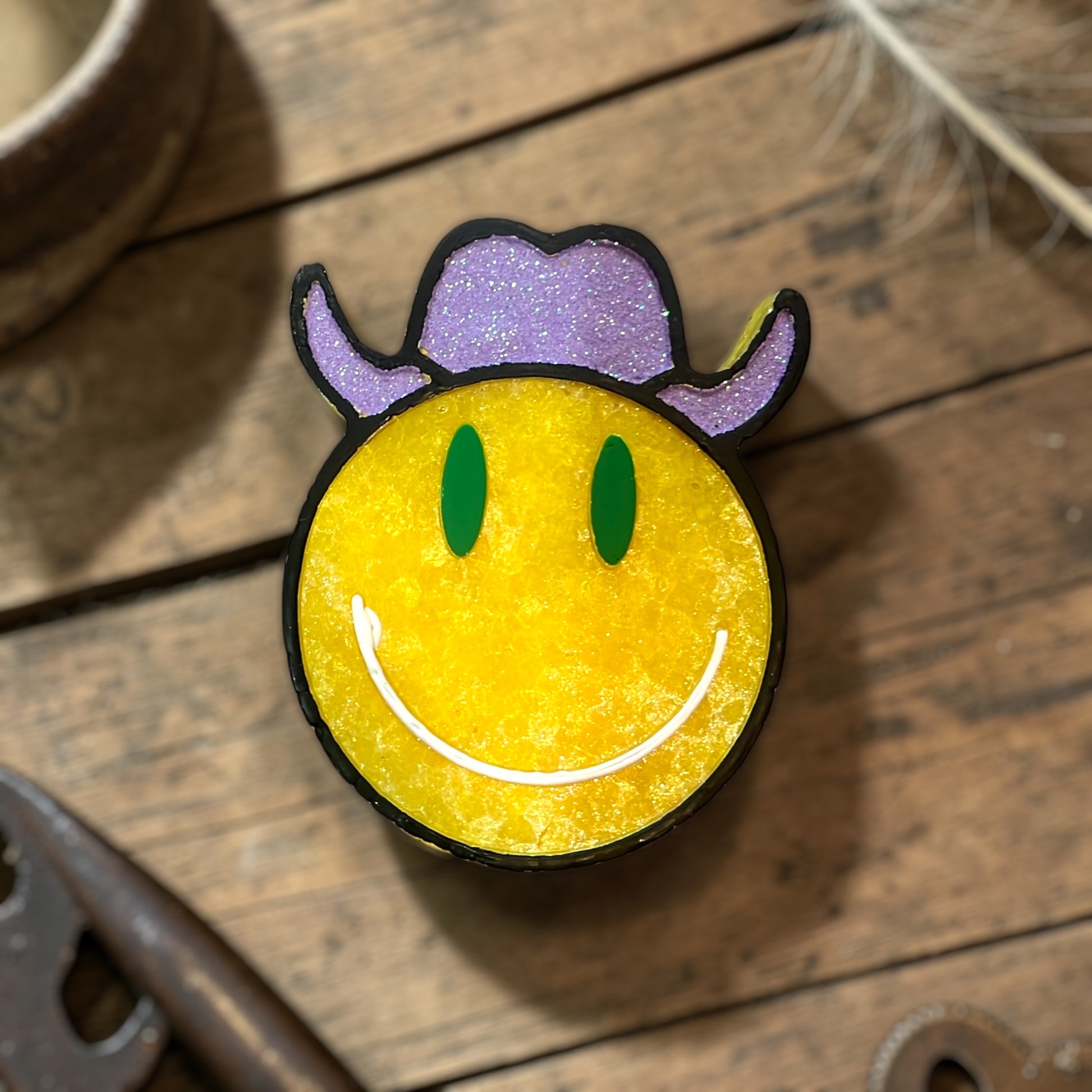 Bright yellow smiley face wearing a purple glitter cowboy hat. Outlined in black with green eyes. On a wooden surface surrounded by western decor.