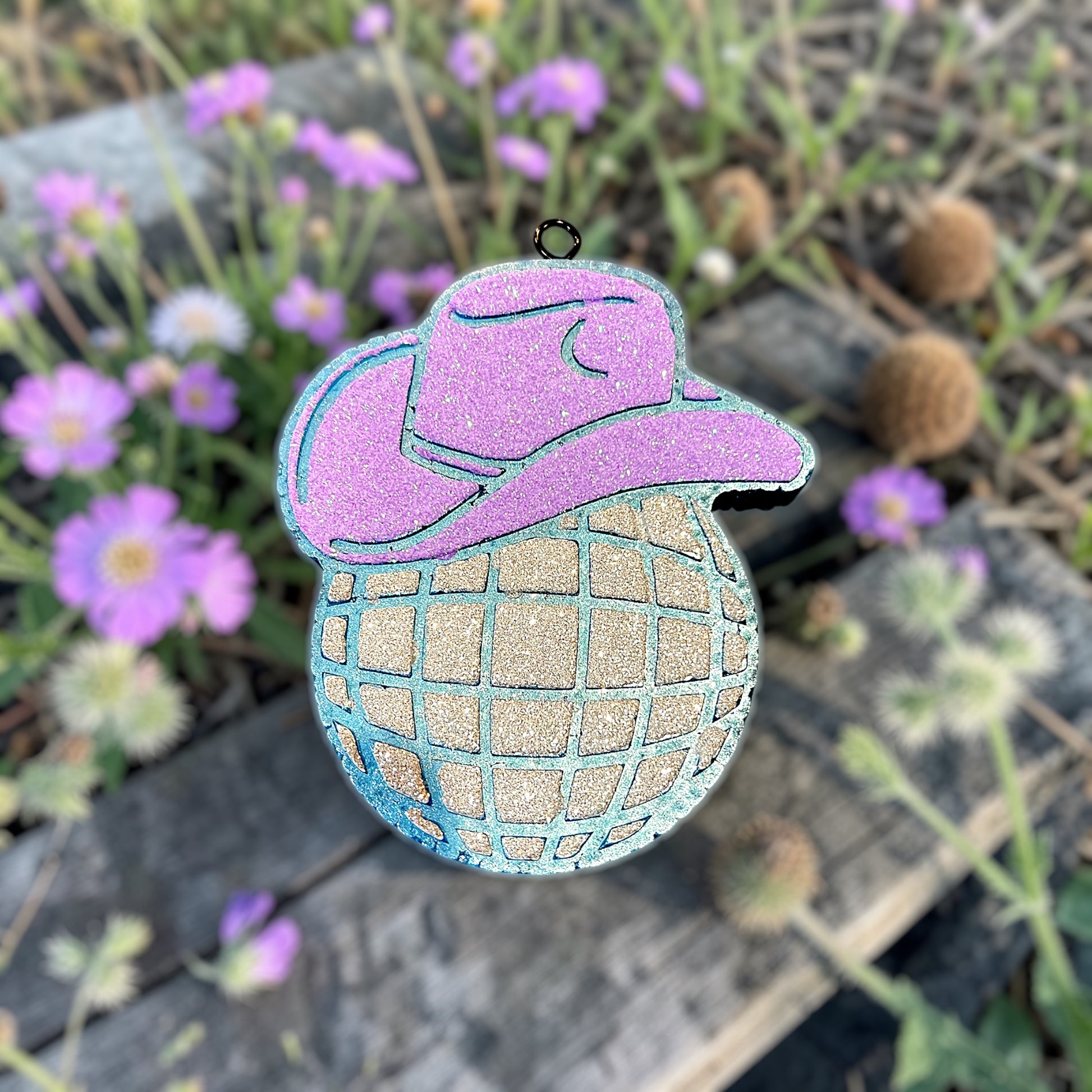 Disco Cowboy car freshie in light blue. Purple glitter cowboy hat and silver glitter disco ball. On a wood bench surrounded by tiny purple flowers.