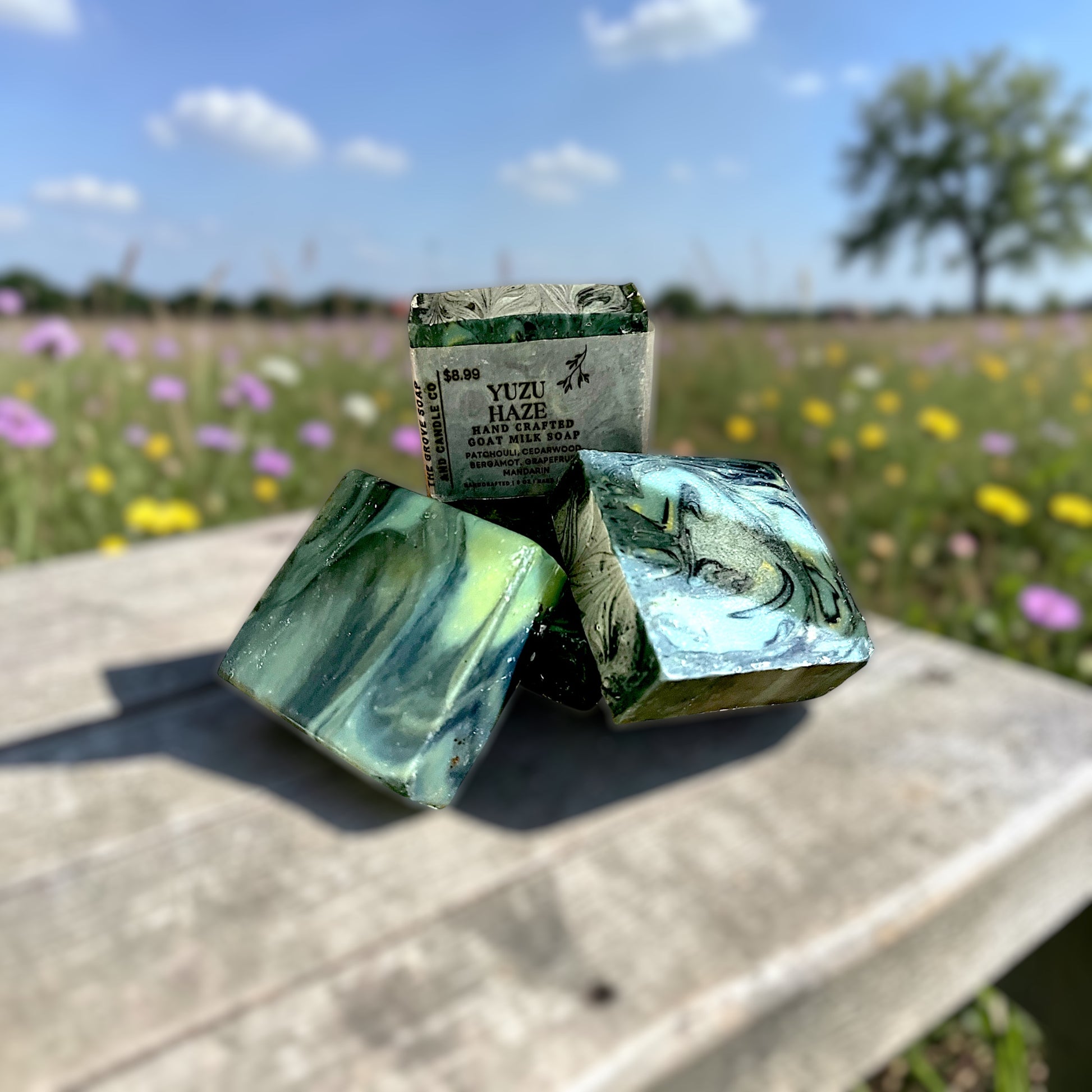 Yuzu Haze goat milk soap on a wooden table with a wildflower field in the background. Green and Yellow swirled bars scented in citrus, bergamot, grapefruit, mandarin, patchouli, and cedarwood.
