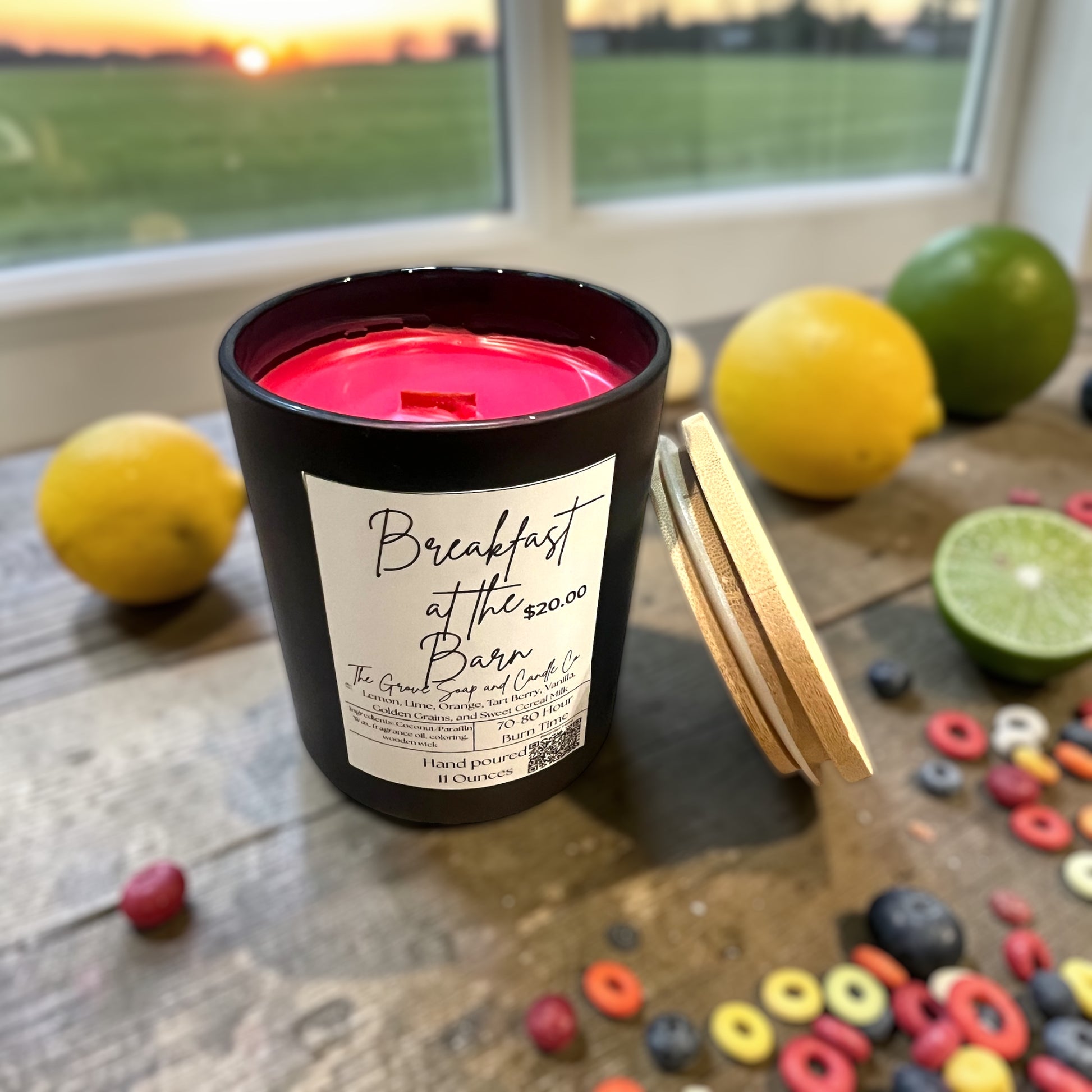 Breakfast at the Barn Candle. A black jar with red wax and a wooden wick. Sitting on a wooden farmhouse table surrounded by lemons, limes, and fruity cereal. 