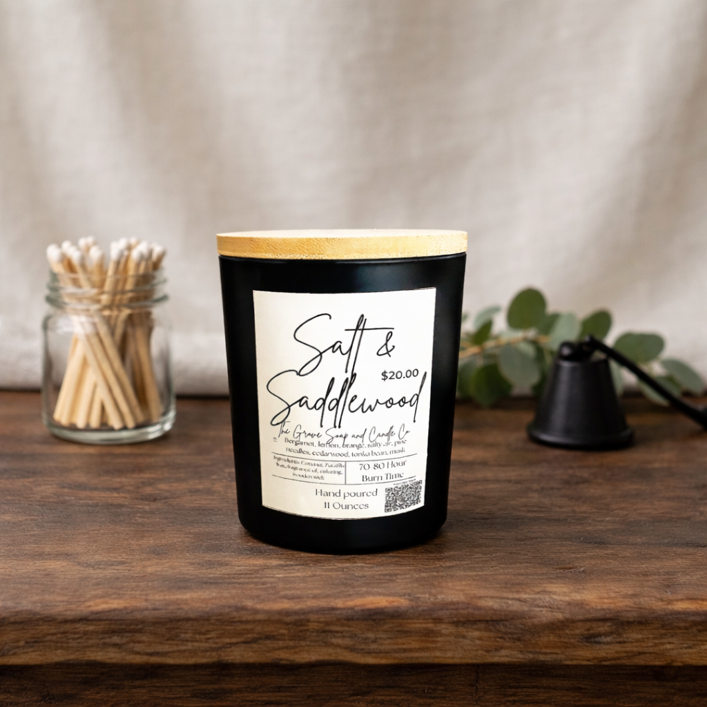 Salt & Saddlewood Candle. A blend of bergamot, lemons, limes, salty air, pine needles, cedar wood, Tonka beans, and musk. Sitting on a rustic wooden table. Matches to the left and a black matte candle snuffed to the right. Background is white linen. 