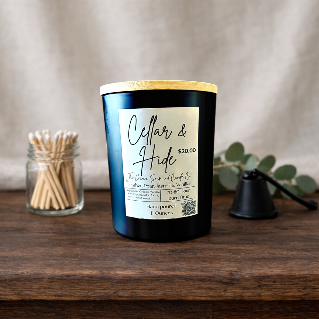 Cellar & Hide Candle a custom exclusive blend of saddle shop leather and sweetened pear champagne. Sitting on a rustic wooden table with a clear bottle of matches to the left and a black matte candle snuffer to the right. A white linen background. 
