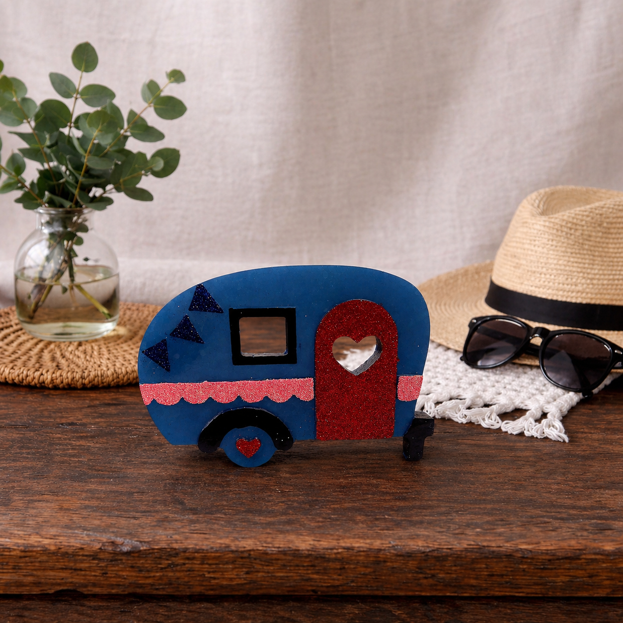 Car Freshie camper on a wooden surface with a hat, sunglasses, and plant in the background.