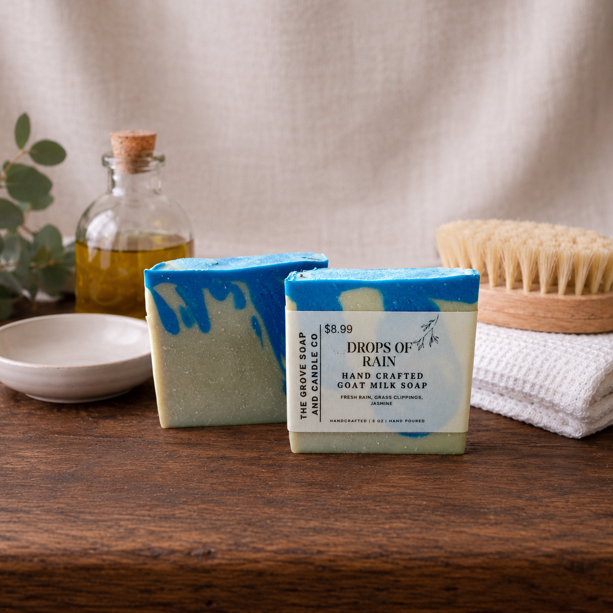 Two bars of soap with a label of Drops of Rain on a wooden surface, surrounded by a bottle, bowl, and towel.