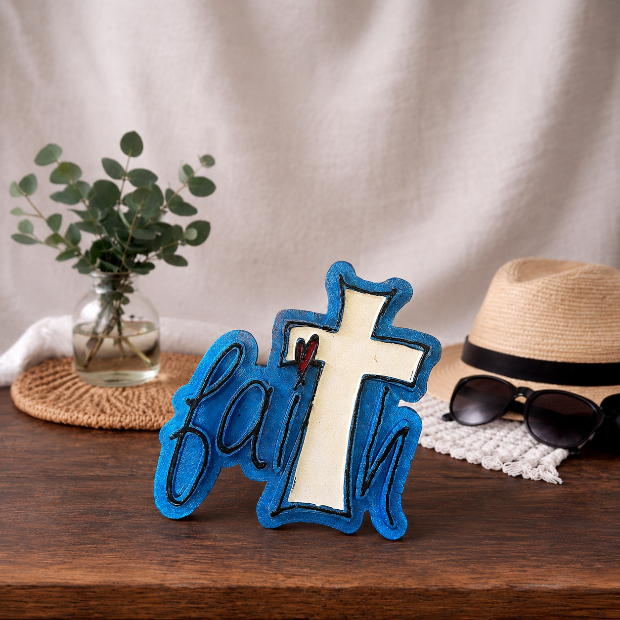 Decorative cross car freshie with 'faith' on a wooden surface with a hat, sunglasses, and plant in the background.
