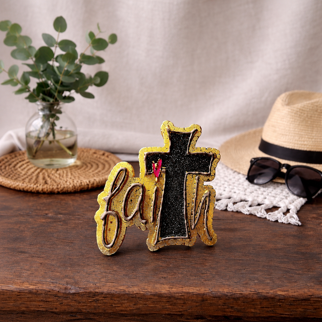 Decorative cross car freshie with 'Faith' on a wooden surface with a hat, sunglasses, and plant in the background.