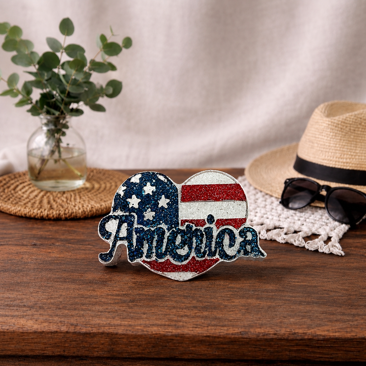 Heart-shaped 'America' car freshie with American flag design on a wooden surface with a hat and sunglasses in the background.