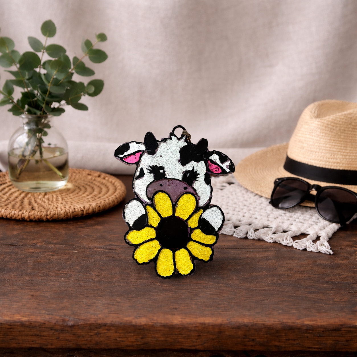 Cow-shaped car freshie with a sunflower design on a wooden surface with a hat, sunglasses, and plant in the background.