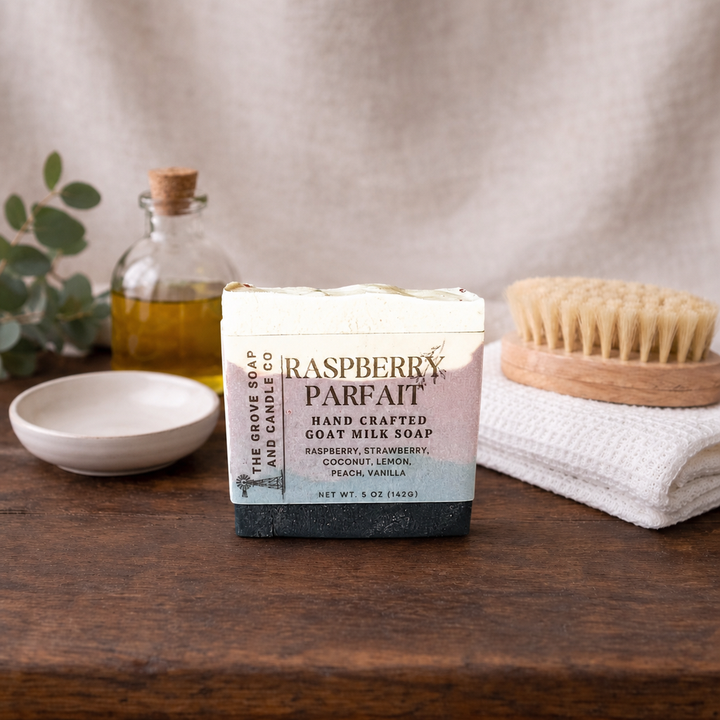Handcrafted goat milk soap bar with packaging on a wooden surface, surrounded by a bottle of oil, a bowl, and a scrubber.