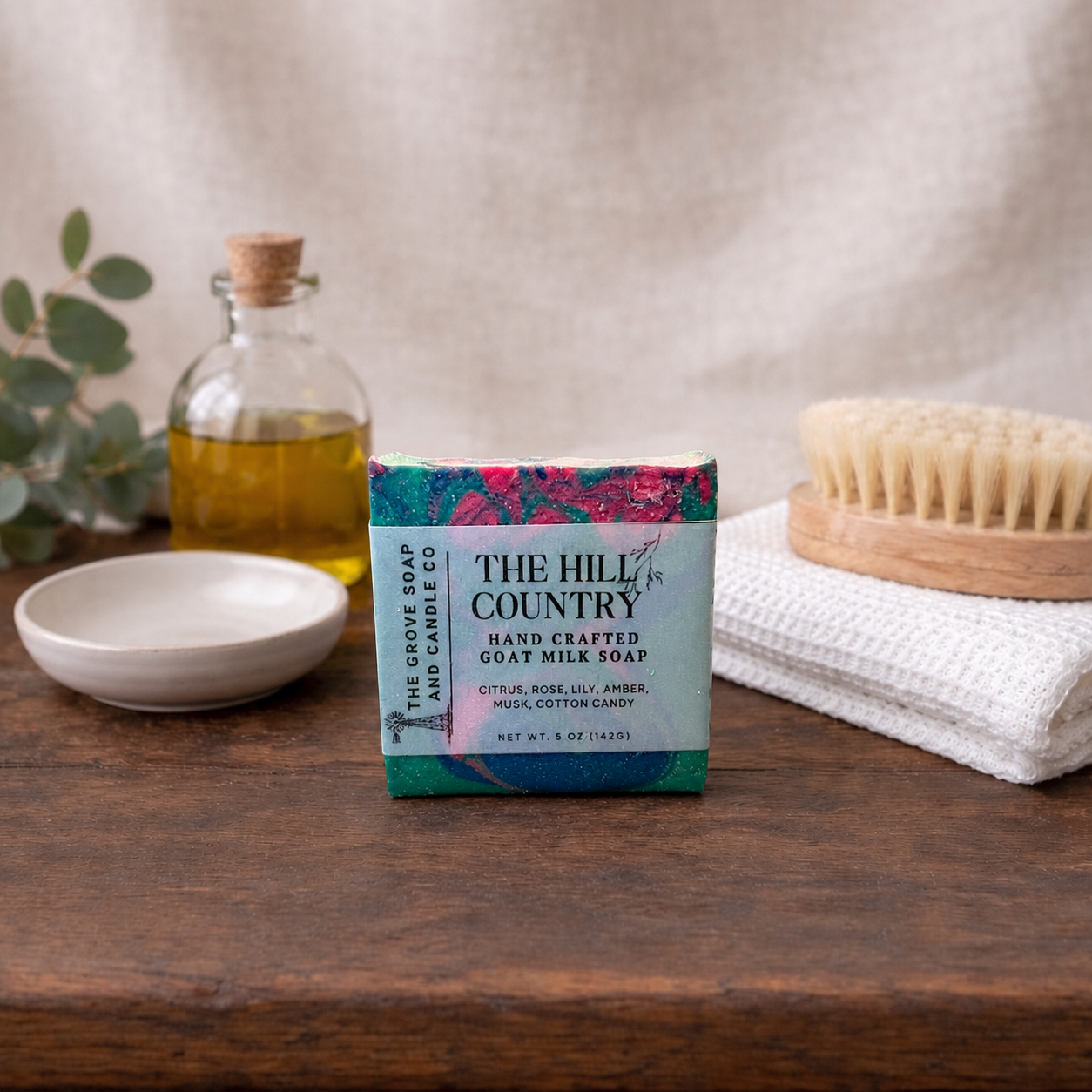 The Hill Country soap bar packaging on a wooden surface with a plant, bottle, and towel in the background.