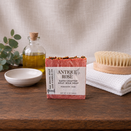 Bar of soap with packaging labeled 'Antique Rose' on a wooden surface with a towel, brush, and oil bottle in the background.