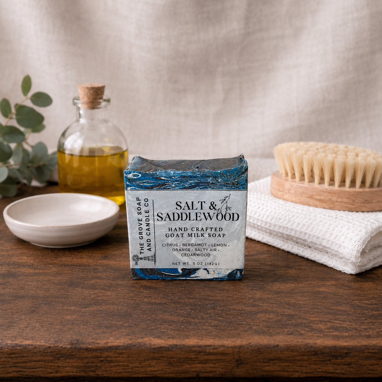 Bar of soap with 'Salt & Saddlewood' branding on a wooden surface with a bottle, towel, and brush in the background.