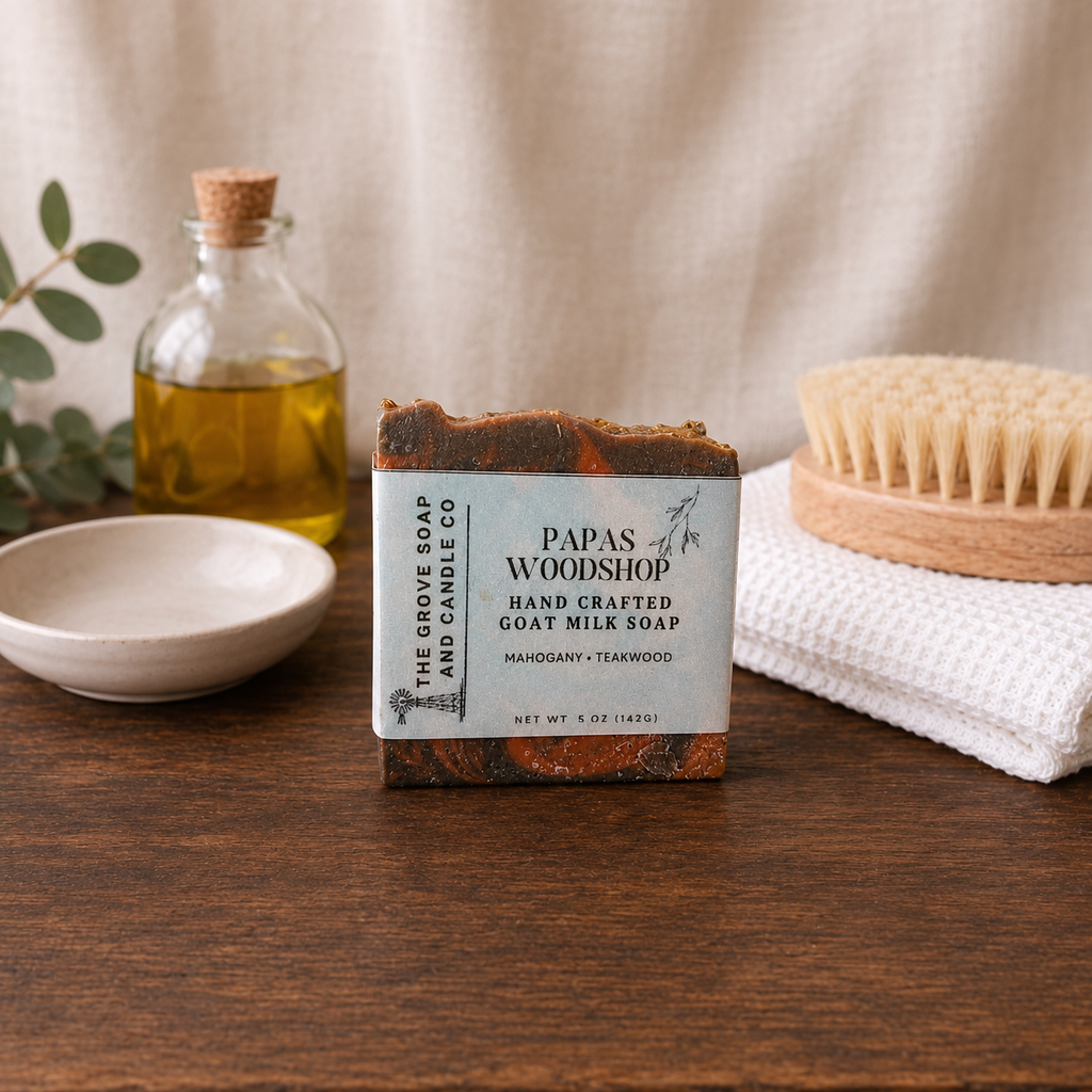 Handcrafted goat milk soap bar from Papas of Woodshop on a wooden surface with a bottle of oil, bowl, and scrub brush.
