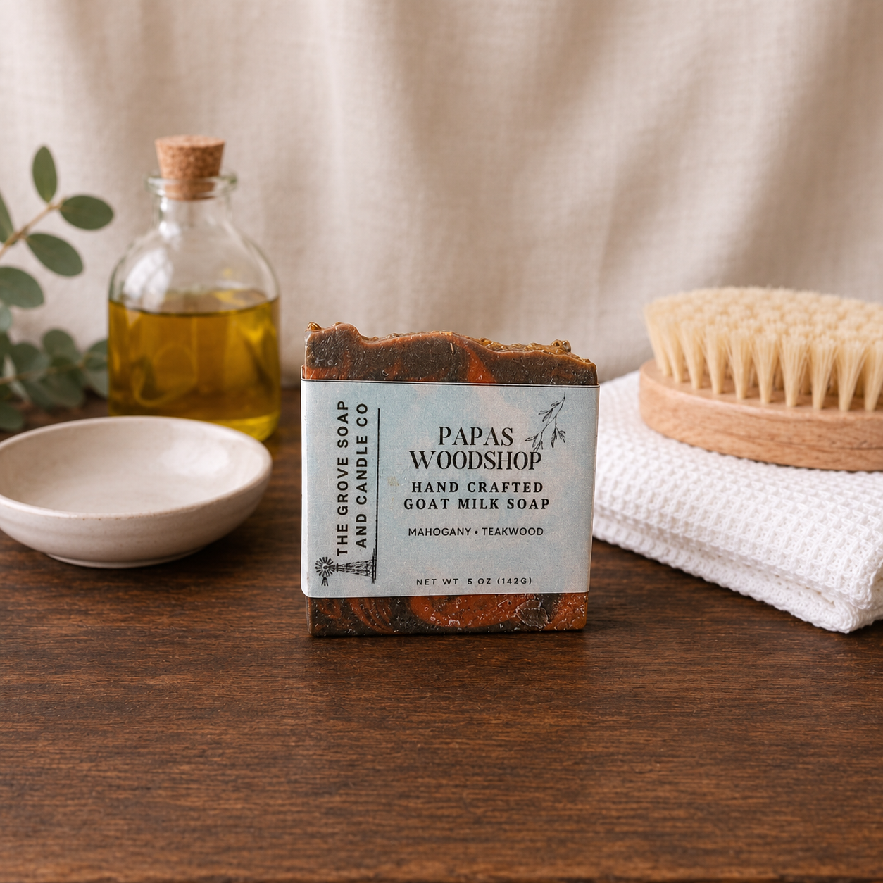 Handcrafted goat milk soap bar from Papas of Woodshop on a wooden surface with a bottle of oil, bowl, and scrub brush.