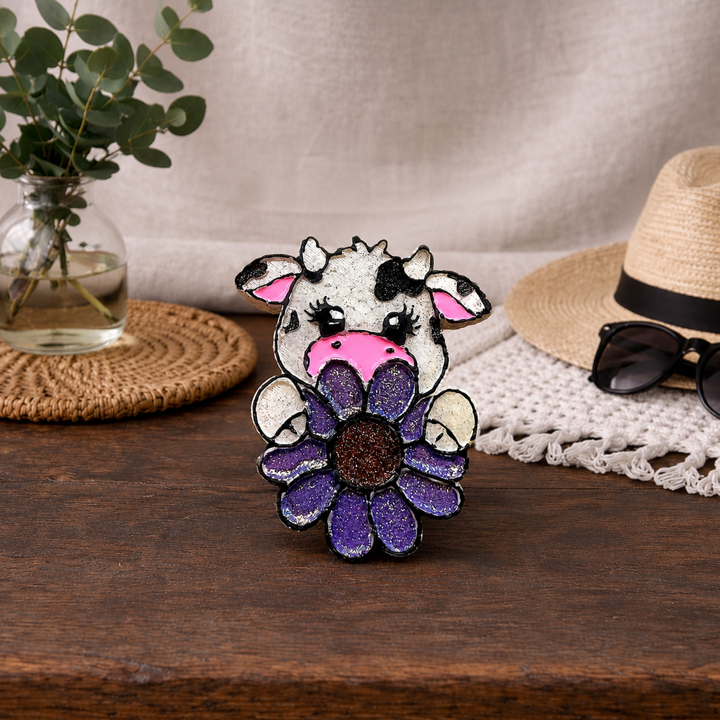 A small decorative cow car freshie charm a purple flower, displayed on a wooden surface with a straw hat, sunglasses, woven placemat, and eucalyptus in the background.