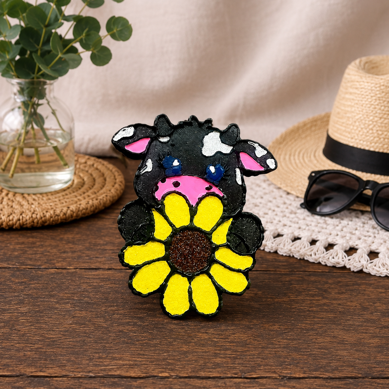 A hand-painted cow car freshie holding a yellow sunflower, displayed on a wooden table with a straw hat, sunglasses, and a glass vase in the background.