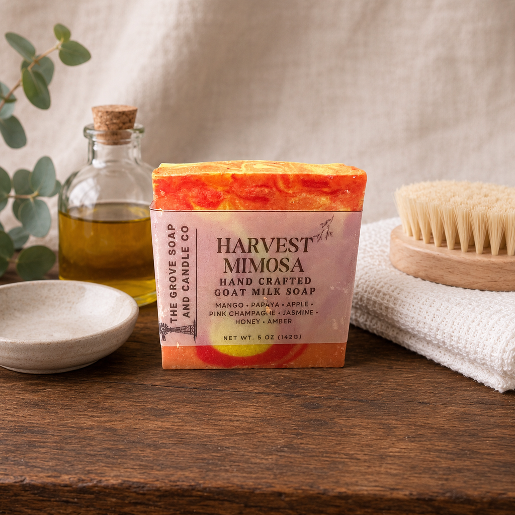 A bar of Harvest Mimosa handcrafted goat milk soap by The Grove Soap and Candle Co. sits on a rustic wooden table, styled with a small bowl, a glass bottle of oil, and a wooden brush against a neutral linen background.