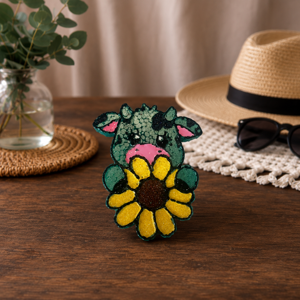 A colorful green and yellow cow car freshie holding a sunflower, placed on a wooden table with a straw hat, sunglasses, and a glass vase in the background.
