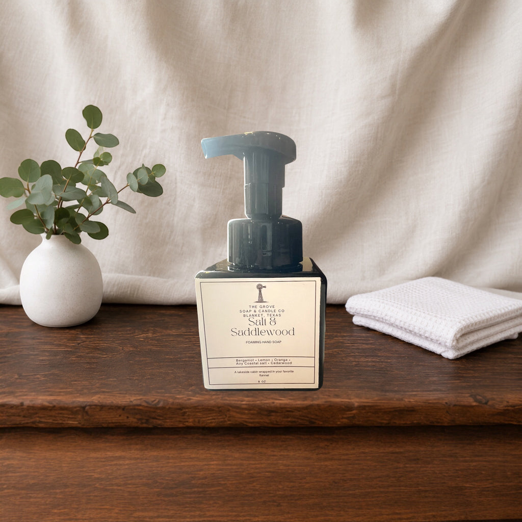 A bottle of Saddlewood Foaming Hand soap with a dark pump and white label, styled on a wooden surface with a small white vase of eucalyptus and a folded white towel against a draped fabric backdrop.