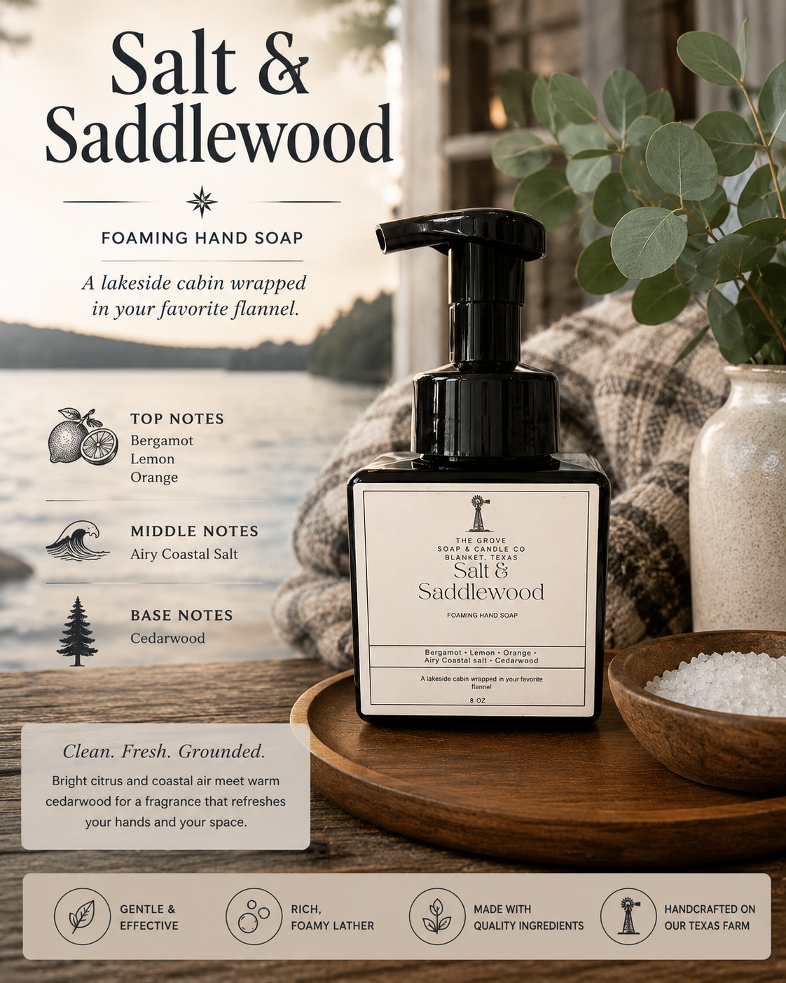 A bottle of Salt & Saddlewood foaming hand soap displayed on a wooden tray in a rustic cabin-style setting with a plaid blanket, bowl of salt, and greenery. The label details the scent notes: bergamot, lemon orange, airy coastal salt, and cedarwood.