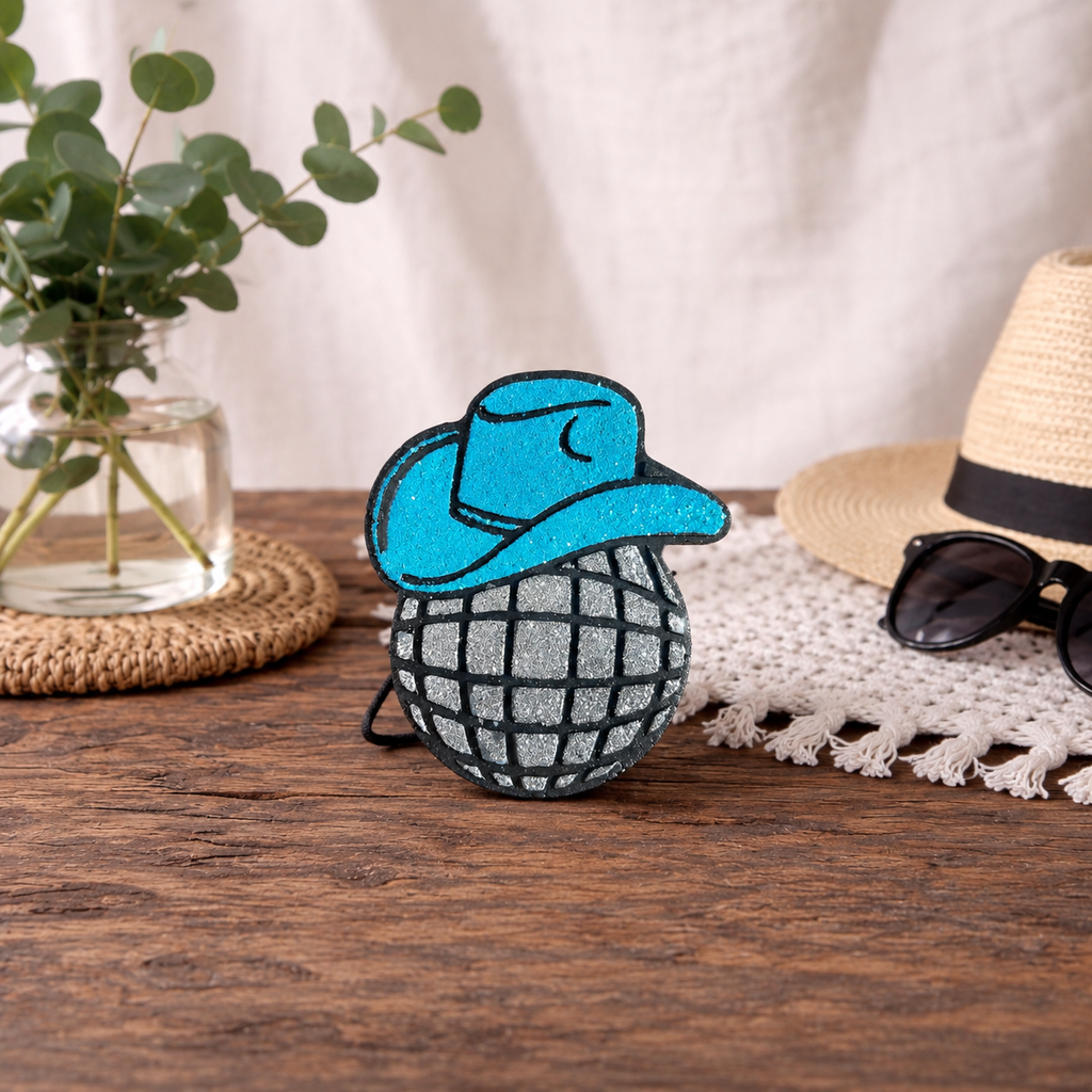 A black and silver glittered disco ball car freshie with a glittered light blue cowboy hat. Sitting on a wooden table. 