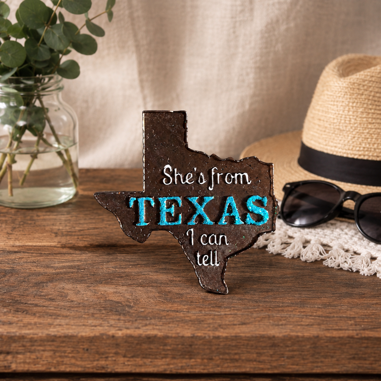 Brown Texas Shaped Car Freshie with light blue glitter that Says Texas. The rest of the car freshie is in white paint and says She's from I can tell. Sitting on a wooden table