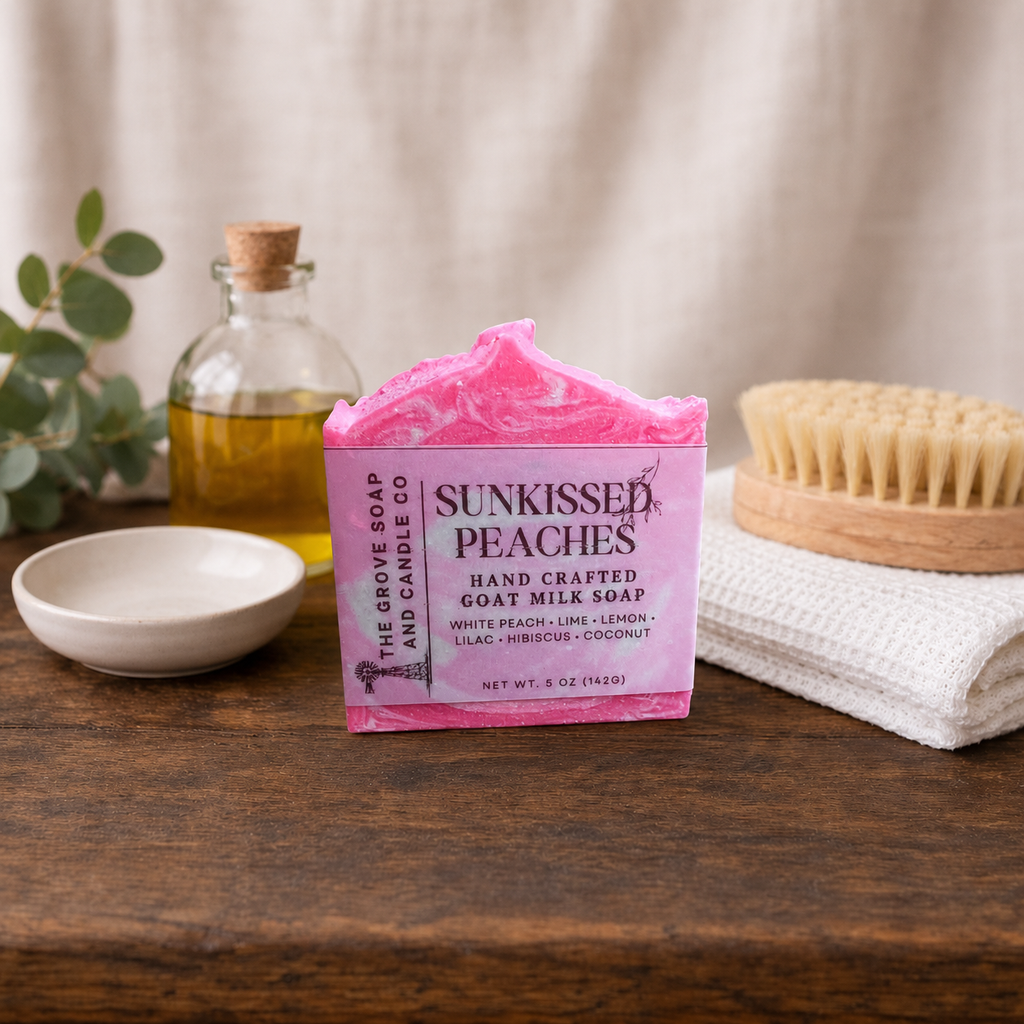 A pink bar of soap labeled 'Sunkissed Peaches' by The Grove Soap and Candle Co sits on a wooden table, styled with a small dish, a glass bottle, a brush, and a folded towel in a warm indoor setting.