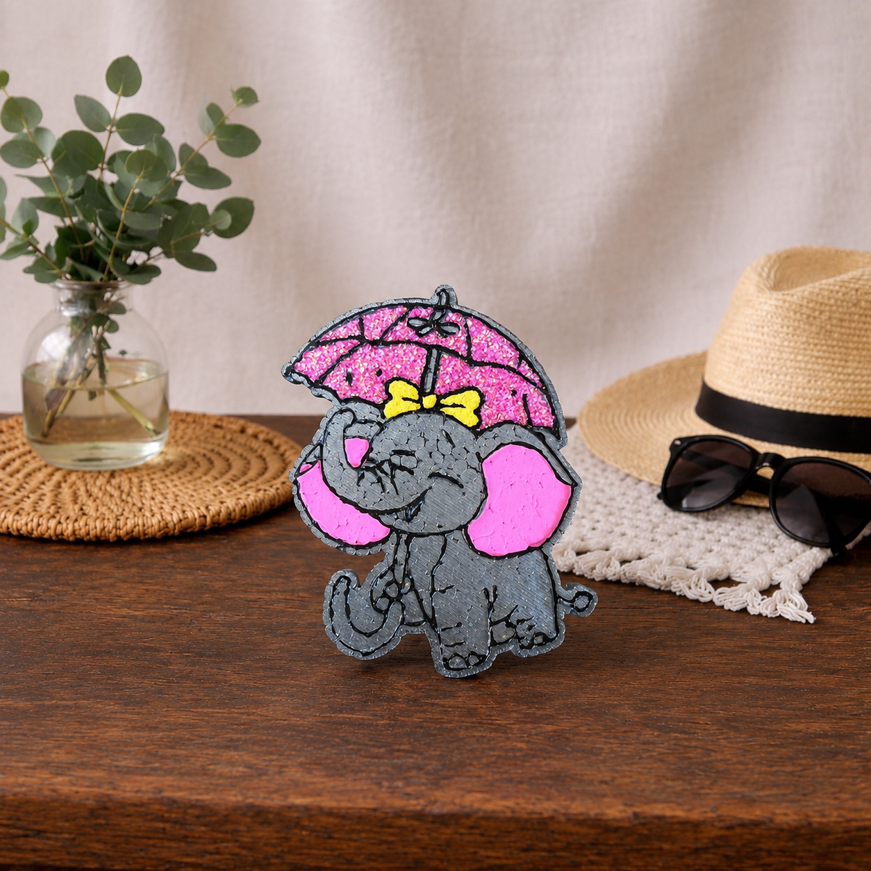Grey elephant car freshie with a pink glitter umbrella and a yellow glitter bow. Sitting on a wooden table. 